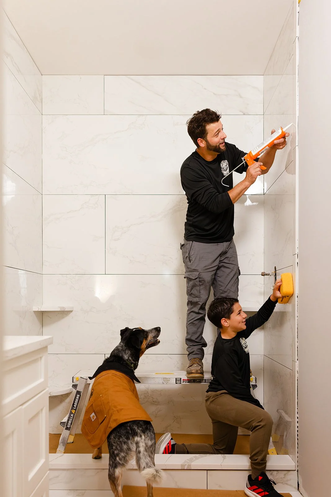 Two men installing tile in a bathroom with a dog watching. One man is on a ladder applying adhesive to the wall, while the other kneels on the floor holding a sponge to clean or smooth the tiles. The dog, wearing a brown jacket, stands nearby observing.