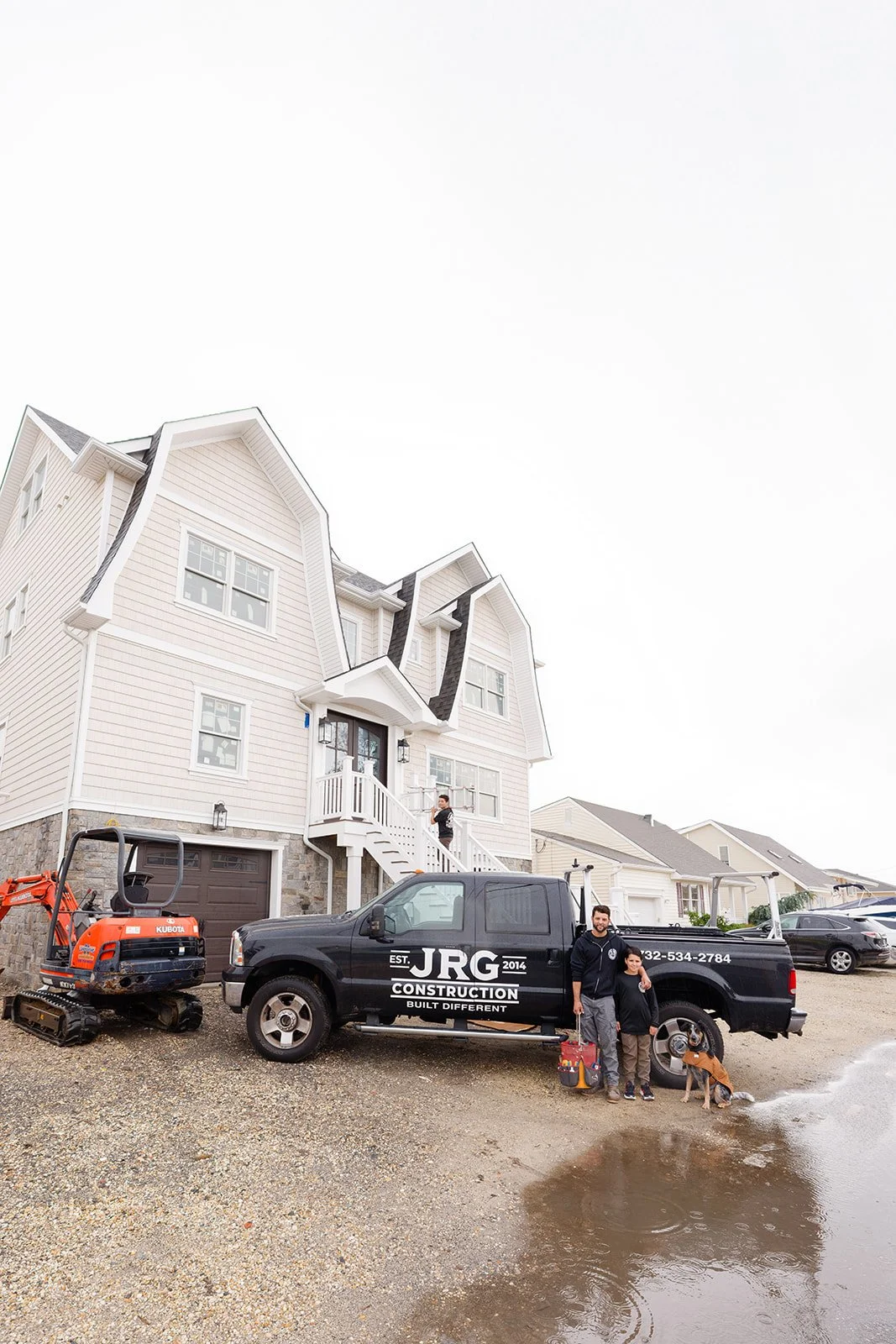 A newly built white house with black roof and large windows, a black construction truck with JRG Construction logo parked in front, a man and boy standing beside the truck, and a small orange excavator on the left. Two other houses are visible in the background.