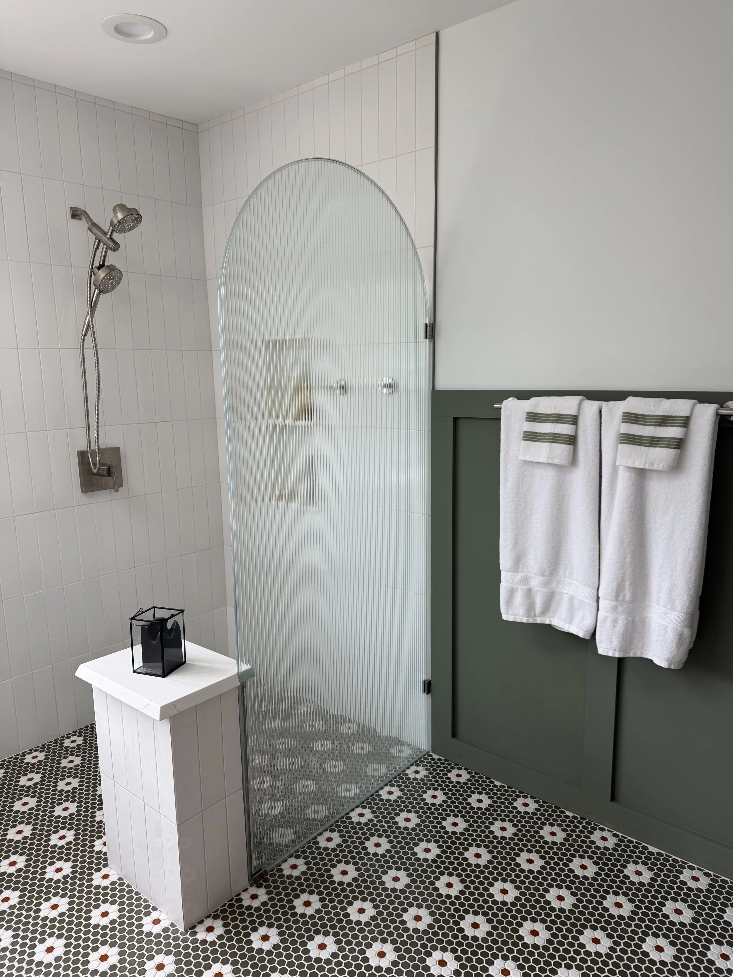 A modern bathroom with a shower area featuring a dual shower head, a white tiled wall, a frosted glass shower partition, a green cabinet with towels, a small white table with a black lantern, and hexagonal mosaic floor tiles with white and reddish-brown accents.