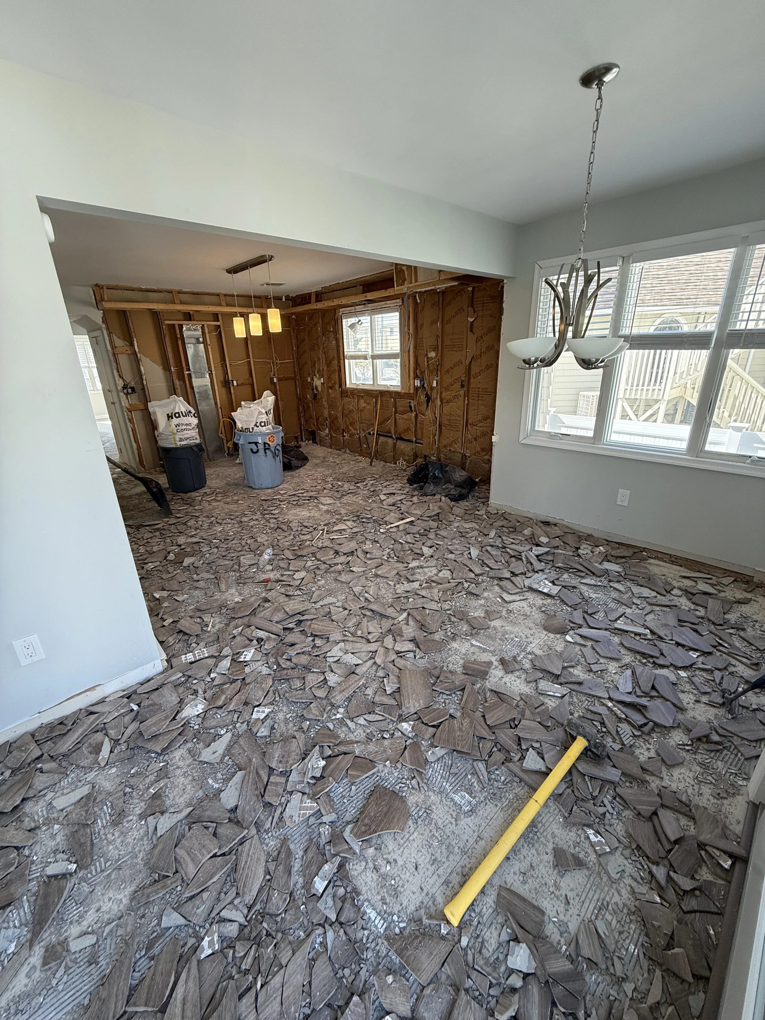 Room under renovation with flooring removed, debris on the floor, dirt, two trash bins, and a construction tool. Part of the wall is exposed showing insulation and framing, with a window and light fixture visible in the background.