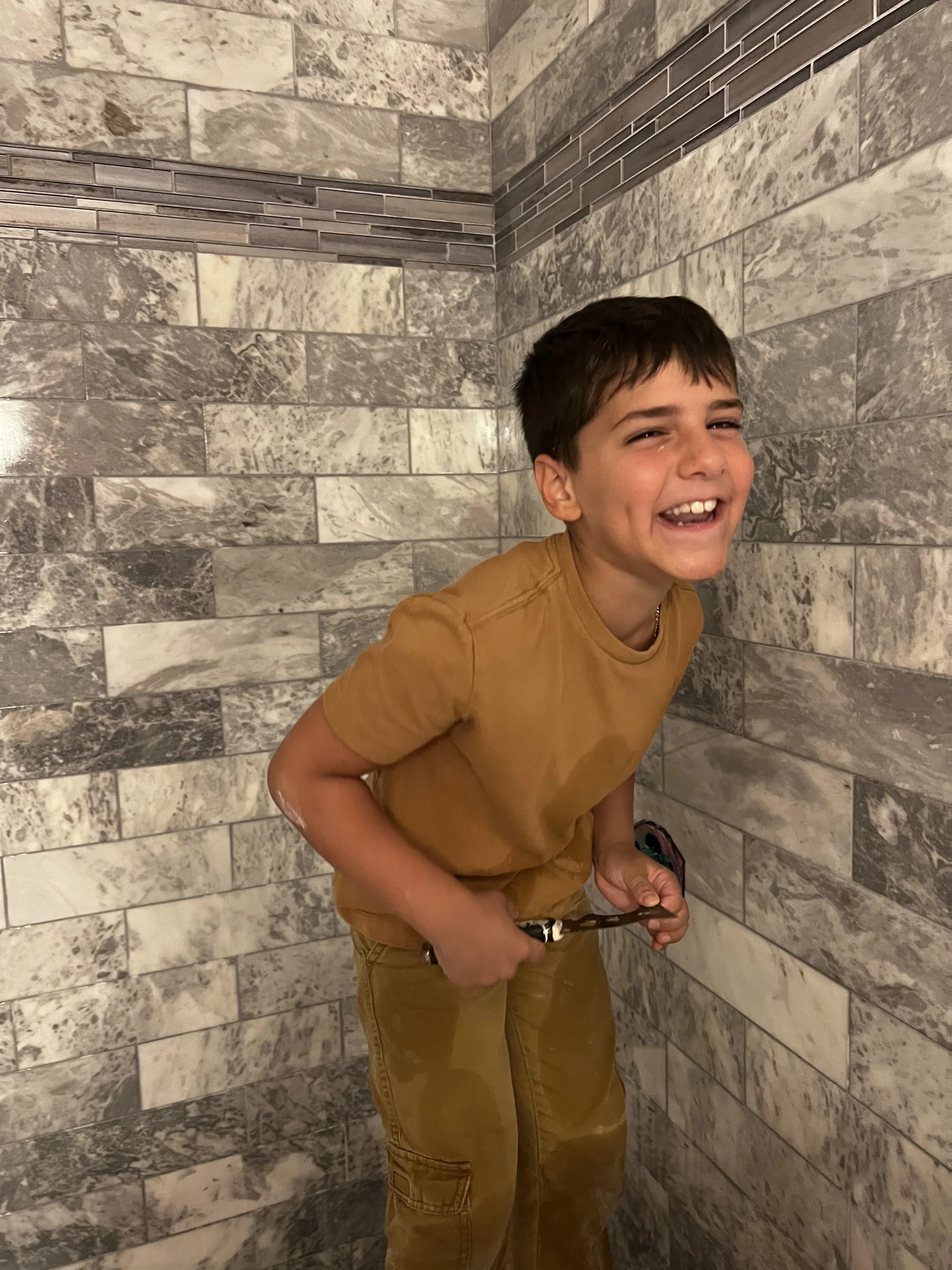 A young boy with short brown hair, smiling and showing teeth, is standing in a corner of a bathroom with grey marble tile walls. He is wearing a brownish-orange t-shirt and tan cargo pants, holding an object in his right hand.