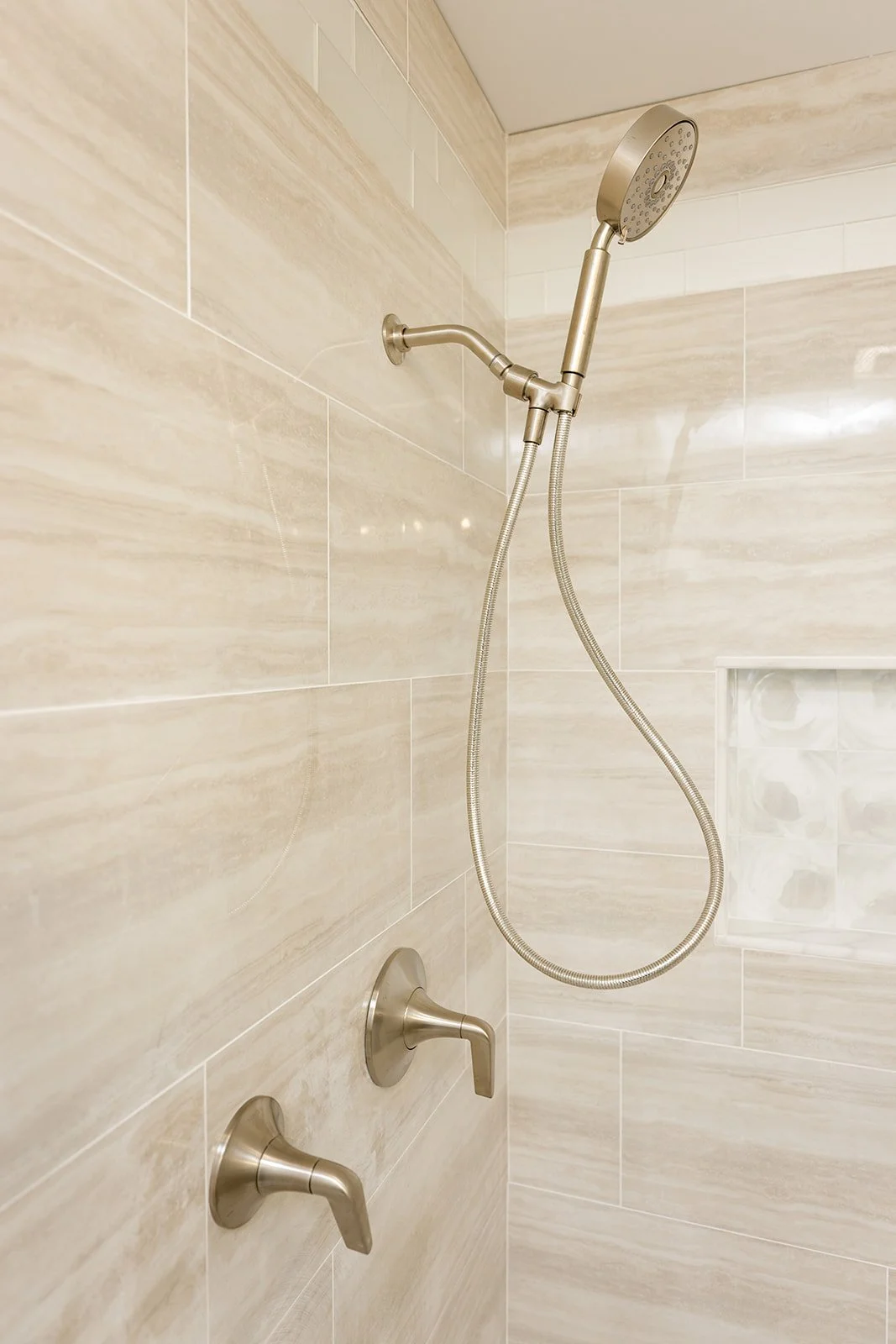 Shower with beige tiled wall, handheld showerhead, and two control knobs.