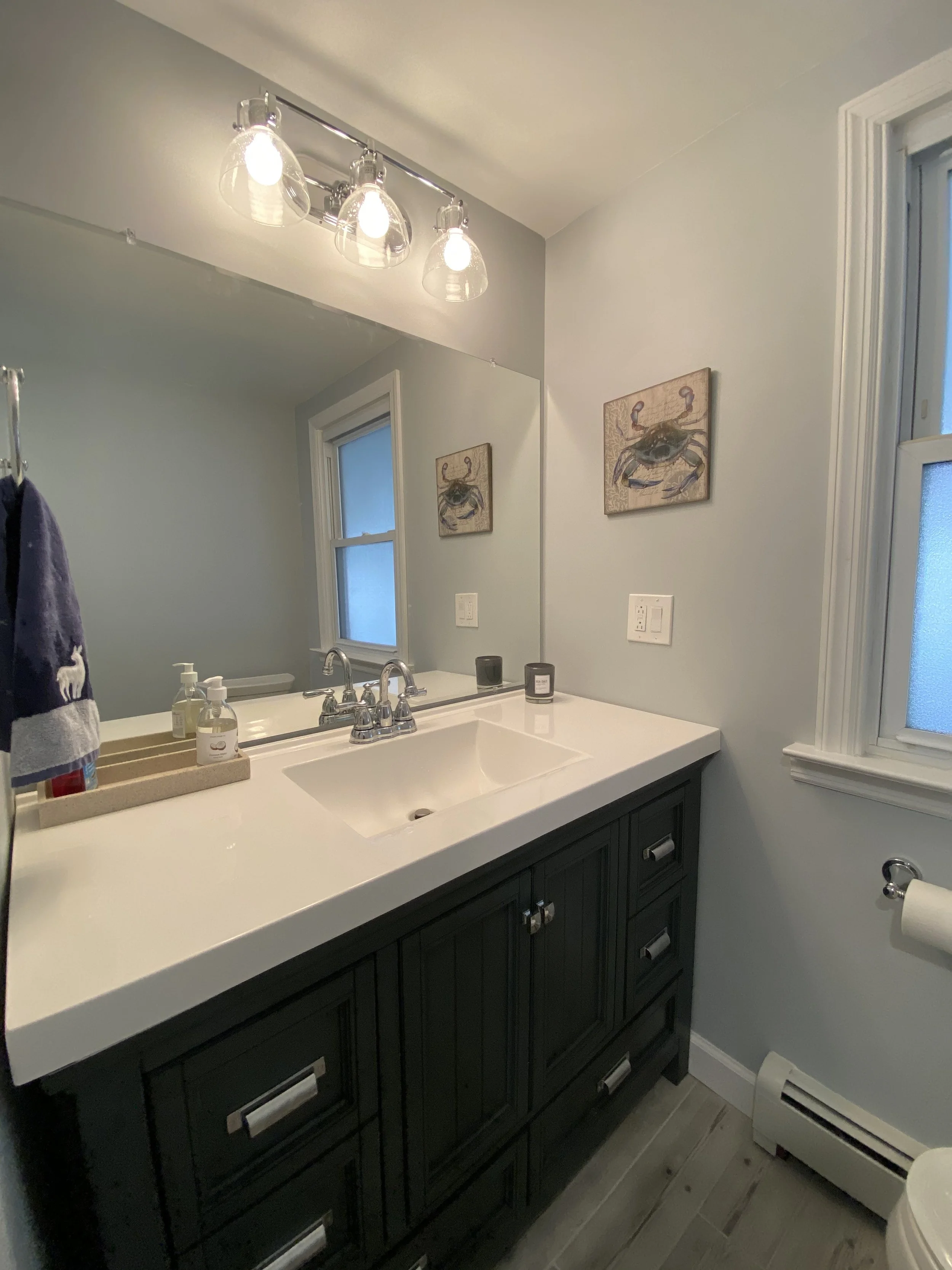 A bathroom vanity with a black cabinet, white countertop, chrome faucet, and a large mirror. There are two candles and a soap dispenser on the counter, and a wall lamp with three glass shades above the mirror. Two small paintings of crabs are hung on the wall, next to a window with frosted glass and white trim.