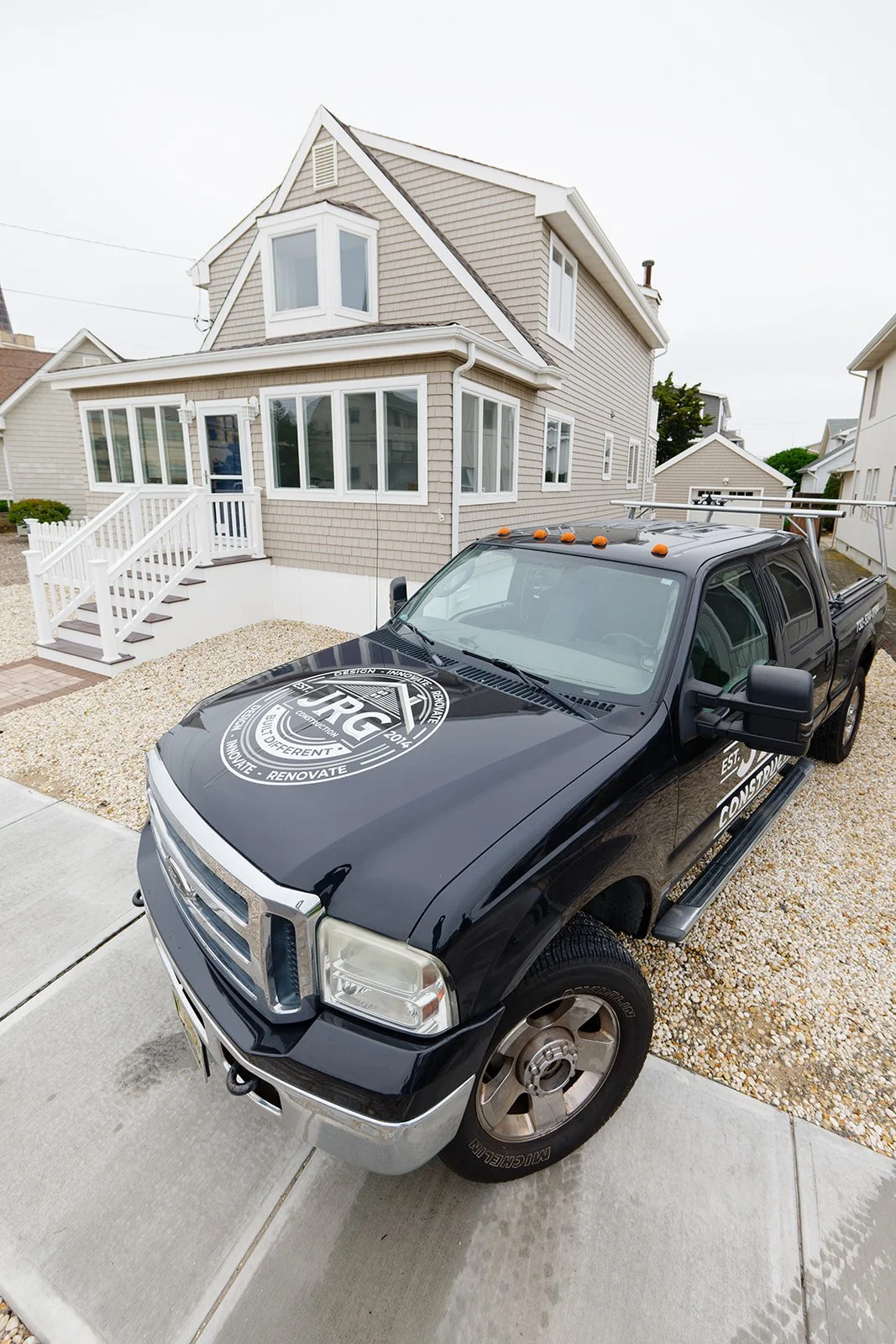 A black pickup truck with company branding parked in front of a two-story house with beige siding and white trim, on a gravel driveway.