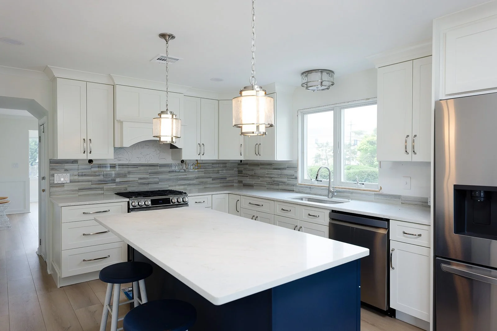 Modern kitchen with white cabinets, gray backsplash, stainless steel appliances, and a white island with navy blue base. There are two pendant lights hanging from the ceiling and a window above the sink.