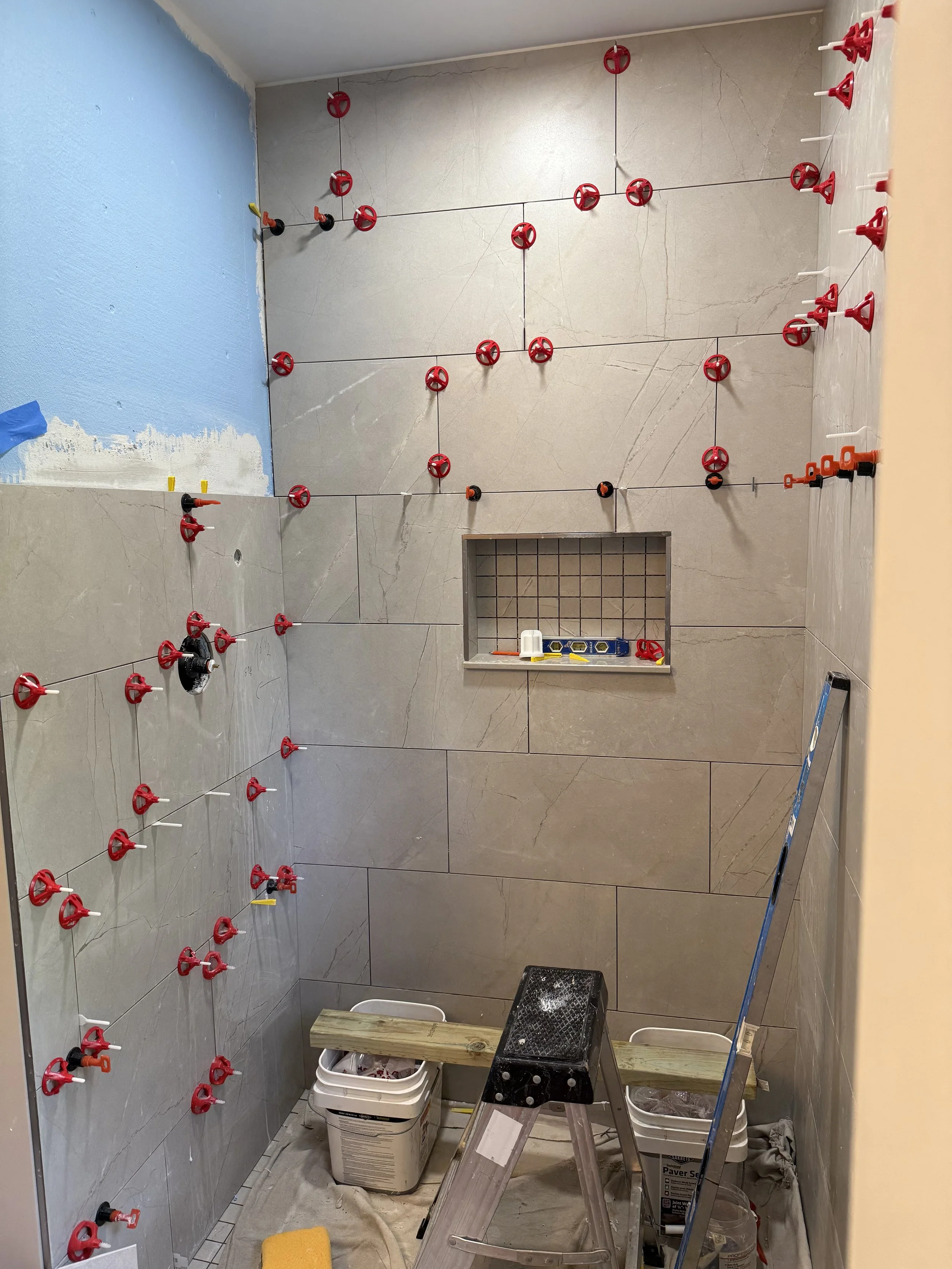 Bathroom under construction with partially tiled walls, red tile spacers on the walls, tools, buckets, a ladder, and a small window.