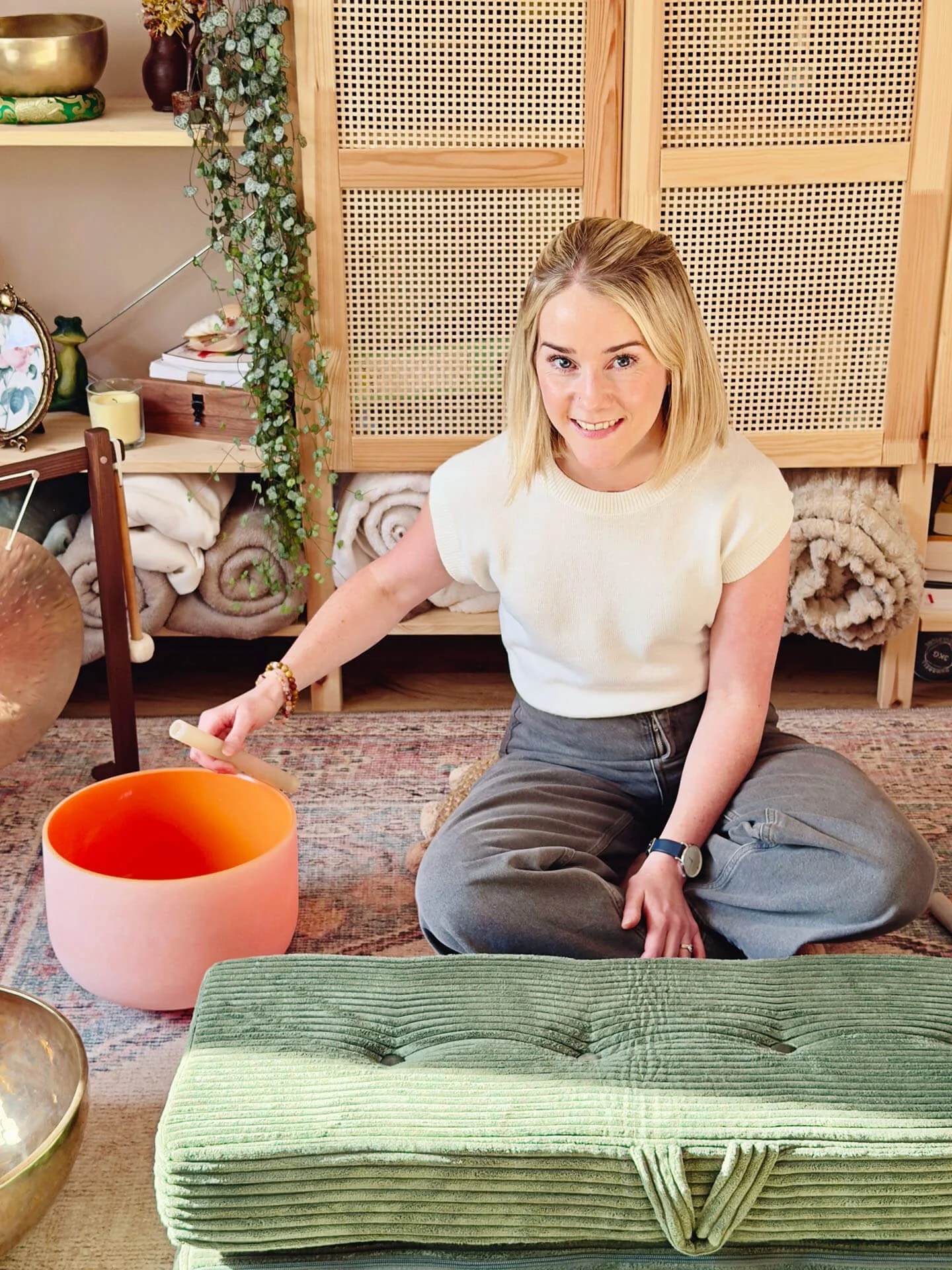 A woman sitting cross-legged on a rug in a cozy room, smiling at the camera, with a singing bowl in front of her and a large green cushion nearby.