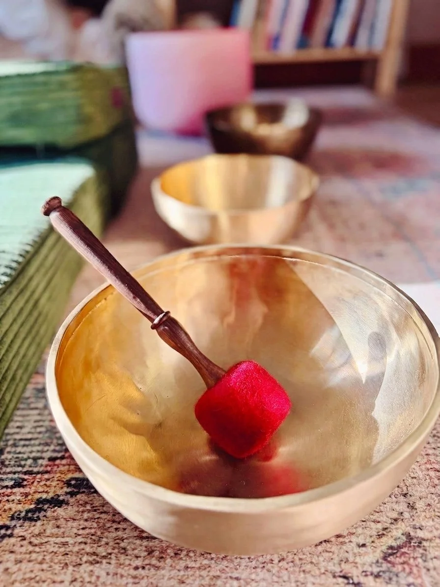 A close-up of a metallic singing bowl with a red mallet resting inside it, situated on a textured surface. In the background, there are other bowls, a pink container, and a bookshelf.