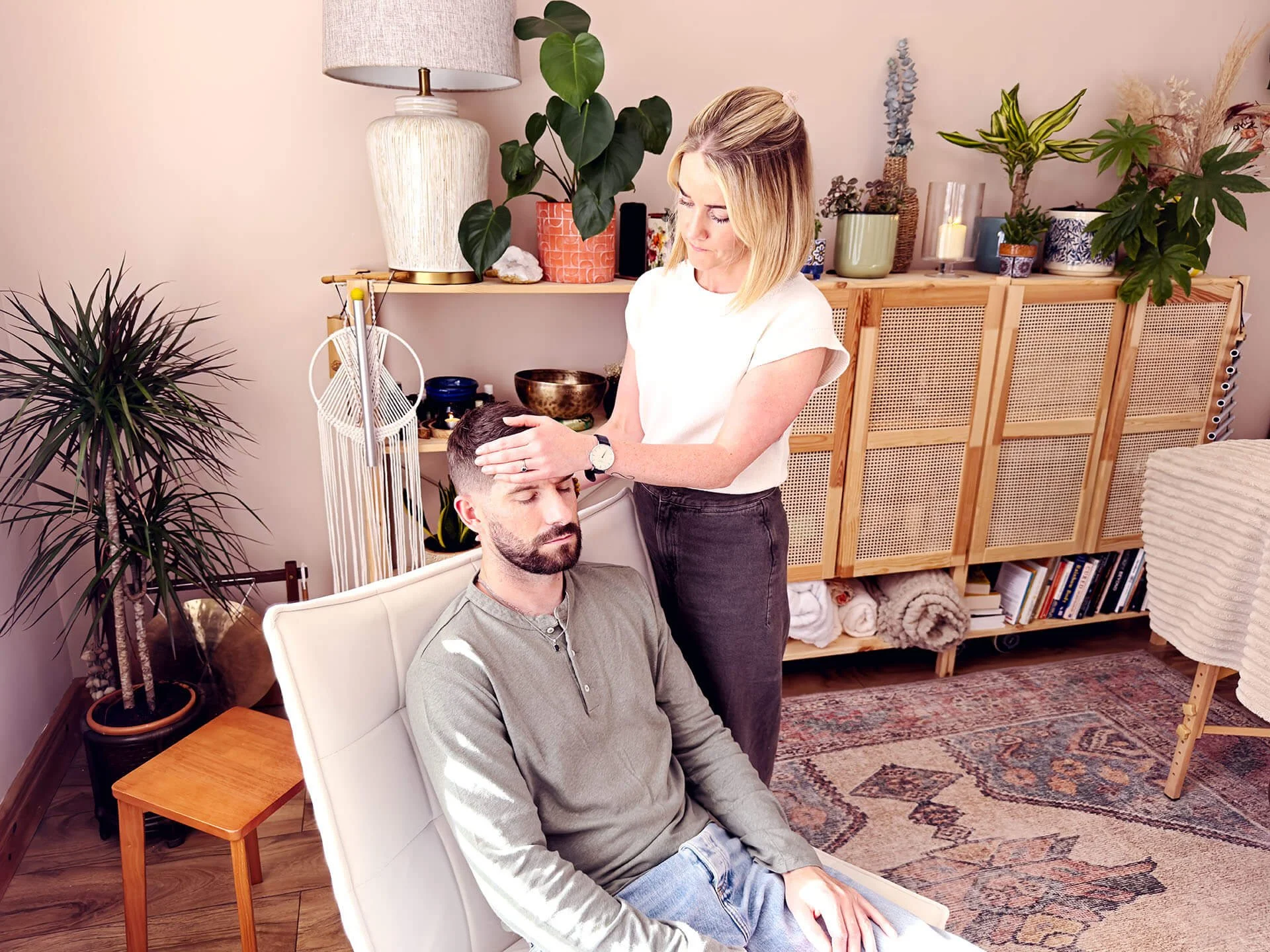 A woman performing energy healing on a man who is sitting with his eyes closed in a comfortable chair in a cozy, well-decorated room with plants, books, and warm lighting.