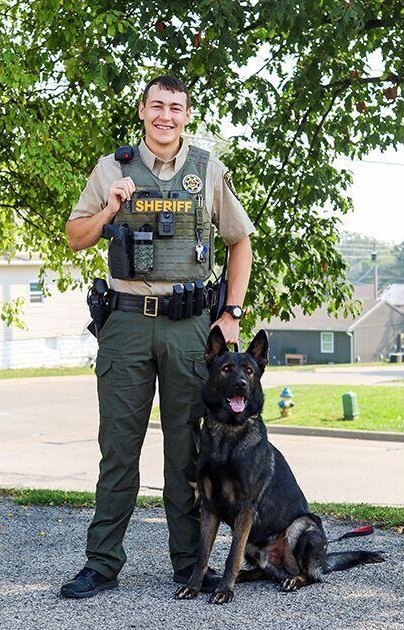 A male sheriff's deputy in uniform standing next to a German Shepherd police dog outdoors with trees and houses in the background.