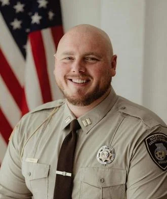 A smiling male police officer in uniform standing in front of an American flag.