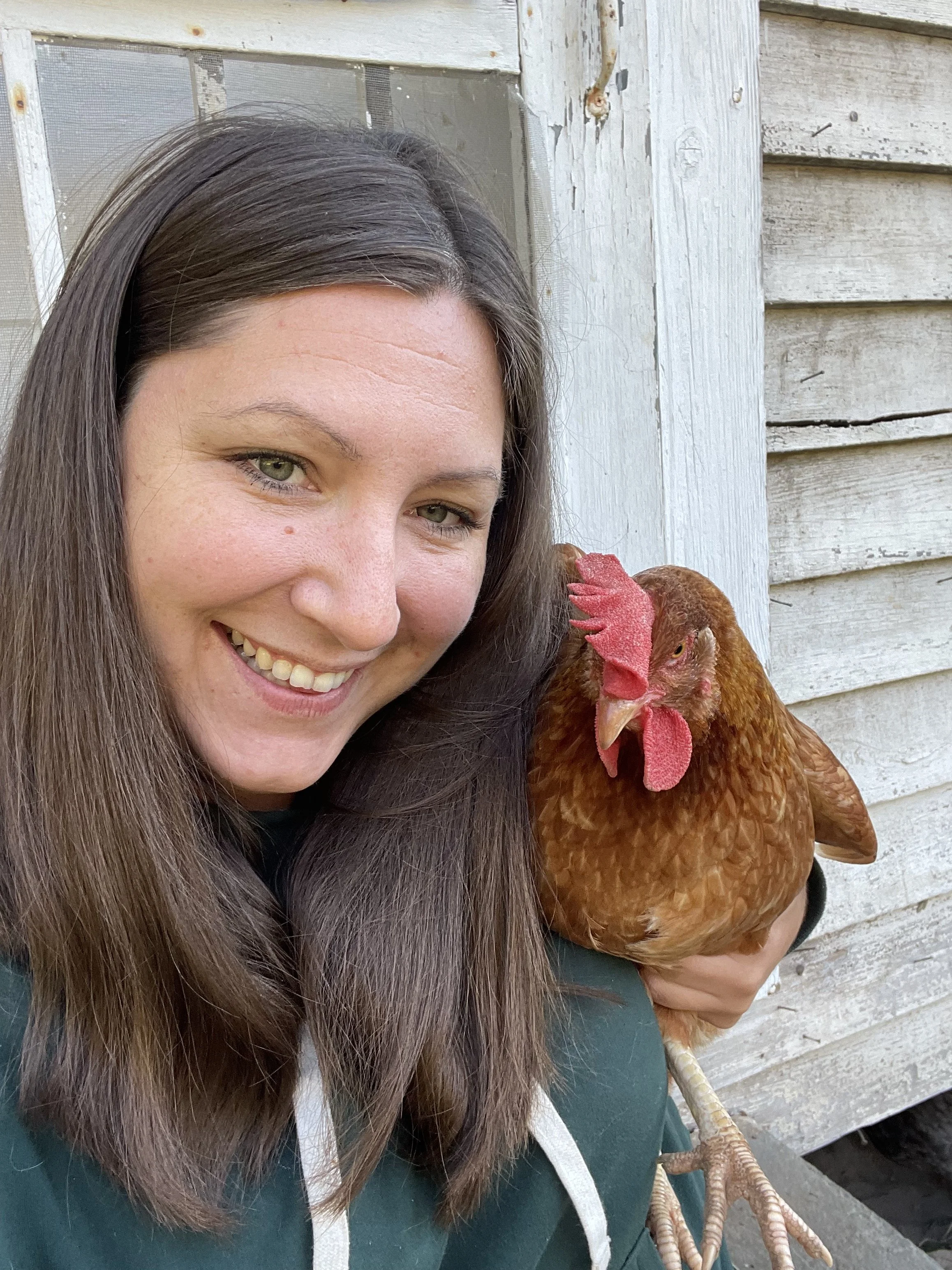A smiling woman with long dark hair holding a brown hen on her arm outside near a wooden building.
