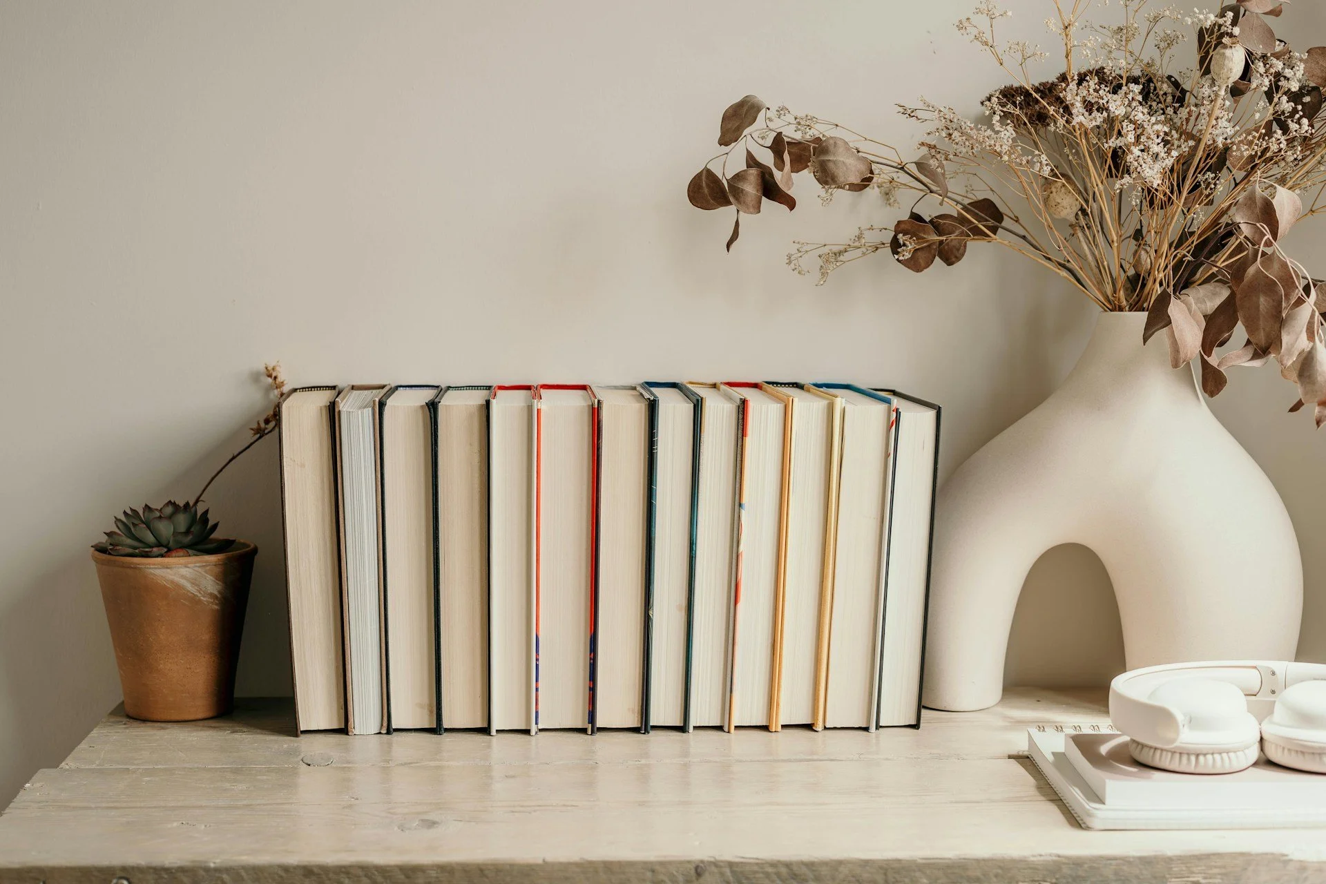 A white bookshelf with a potted succulent, a row of books, and decorative items including a large white vase with dried flowers, a small white bowl, and a white bowl with a lid, all arranged on a light-colored wooden surface.