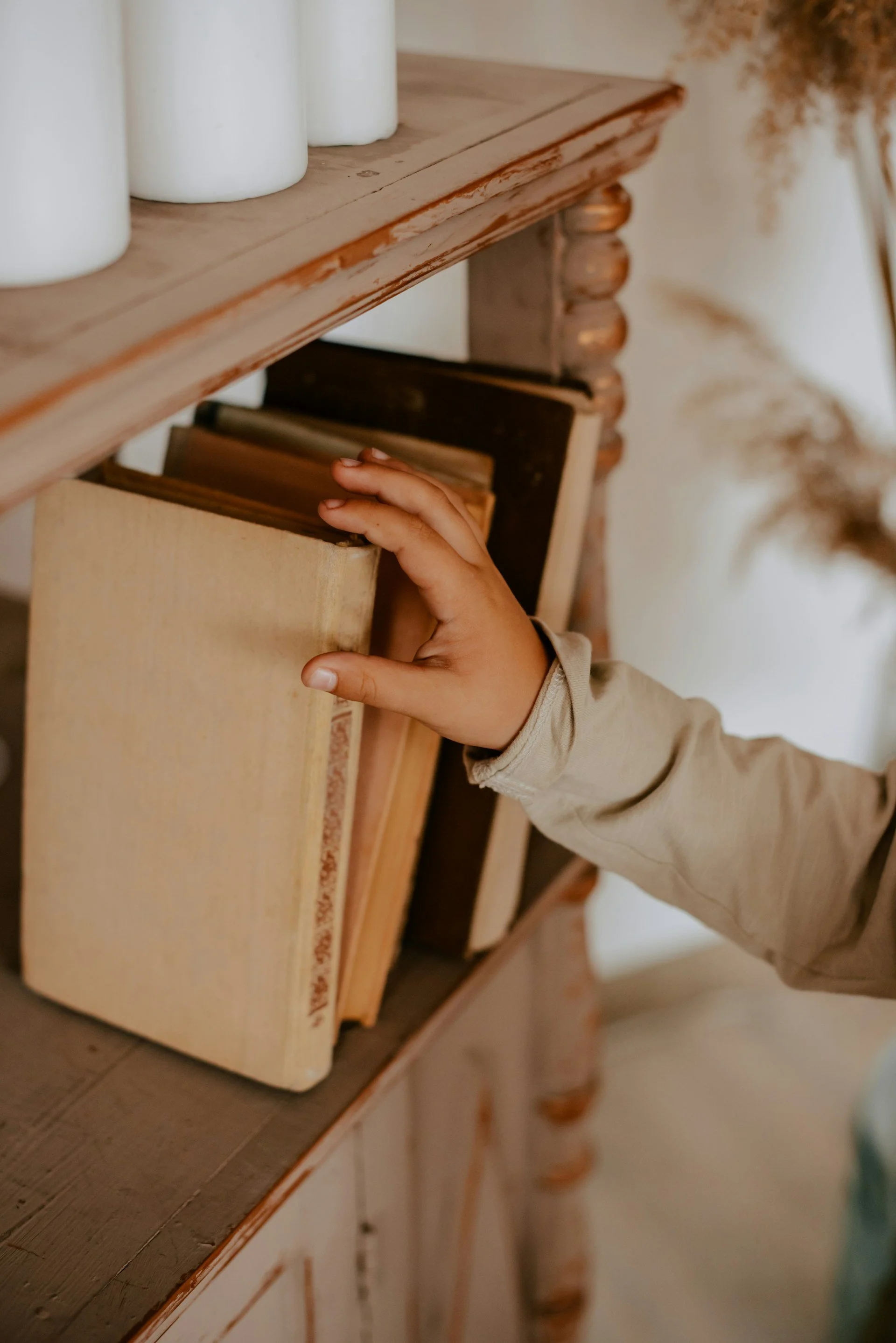 A child's hand reaching into a wooden bookshelf filled with worn books, with a wooden mantle and white ceramic vases on top in the background.
