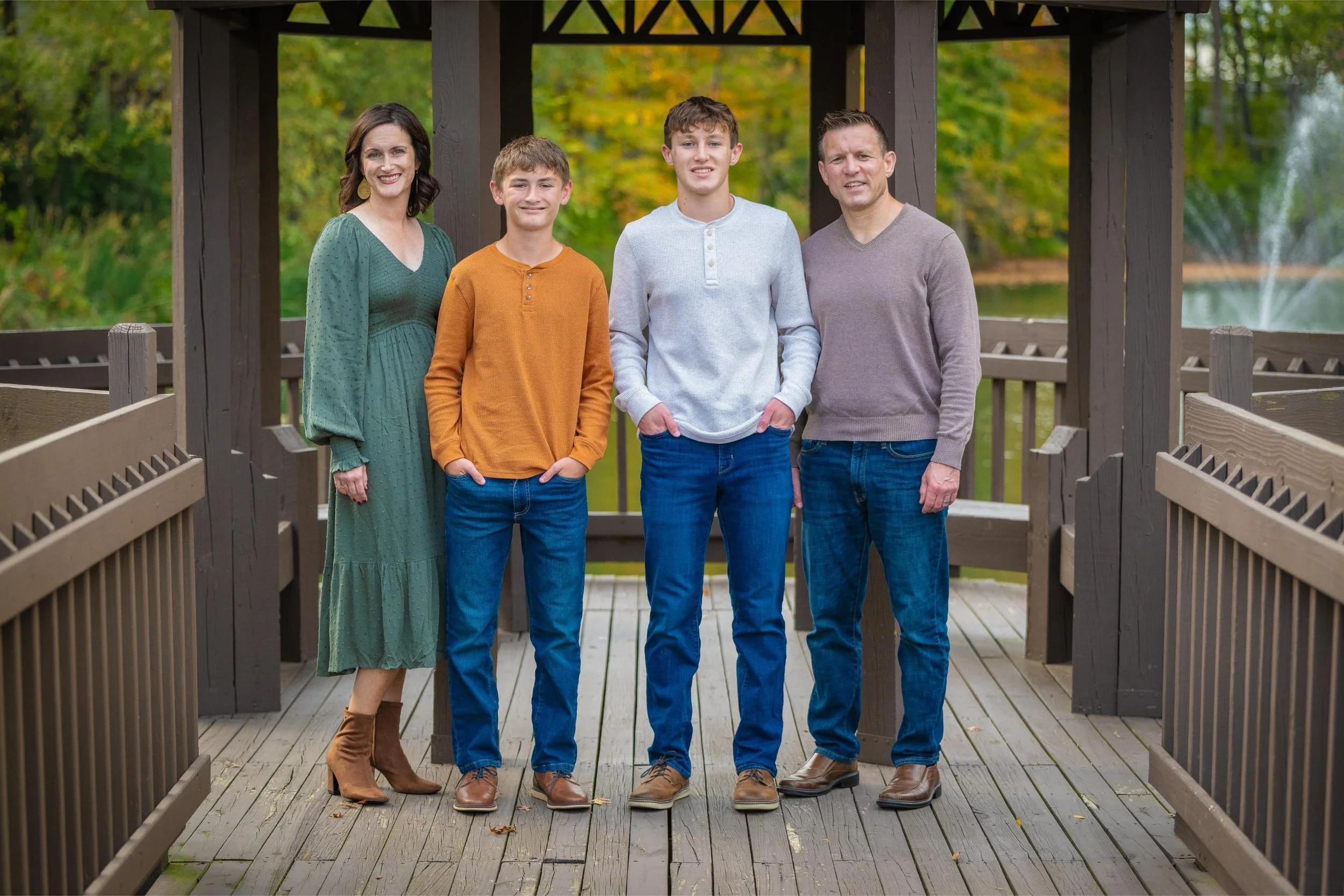 Krista Harrington's family of four posing on a wooden bridge in a park with trees in the background. They are all smiling and dressed casually for fall.