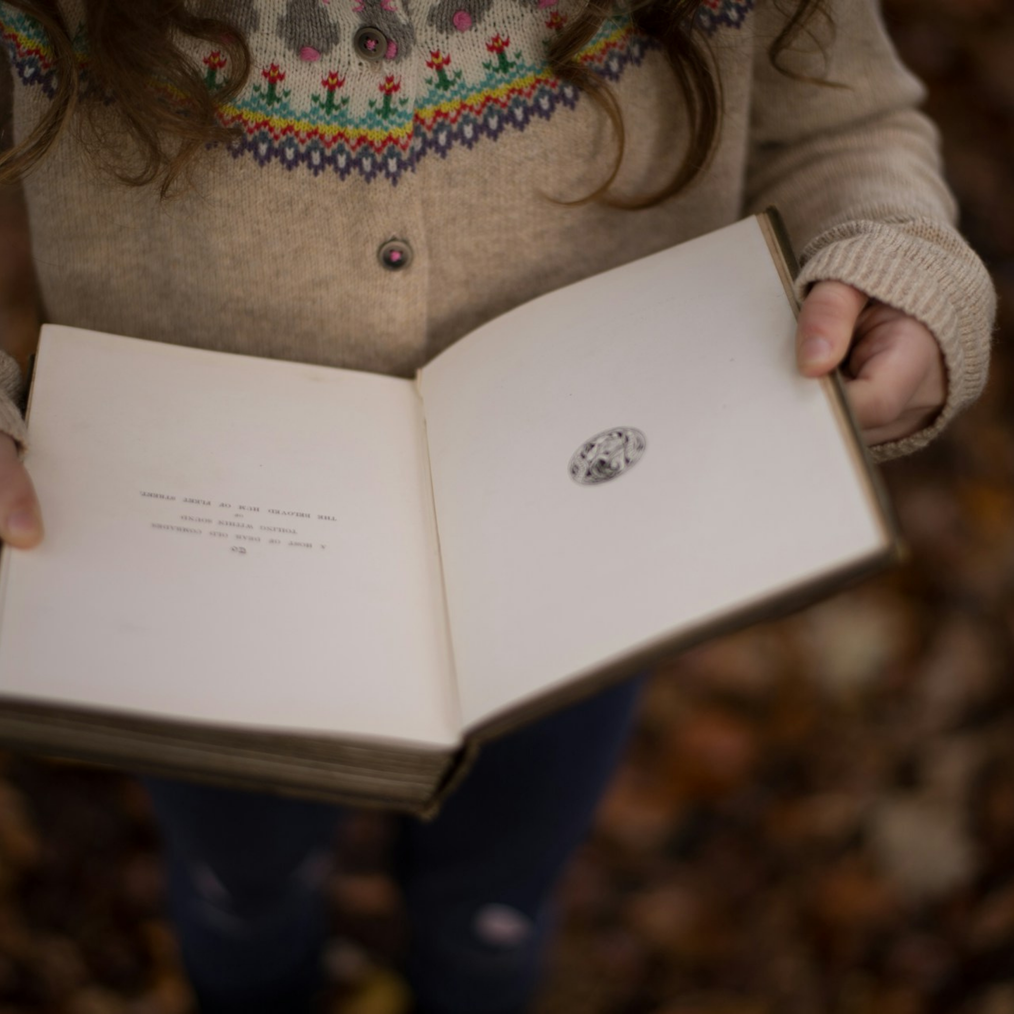 A young child with long, curly hair wearing a beige sweater with colorful, patterned embroidery around the neckline, holding an open book.
