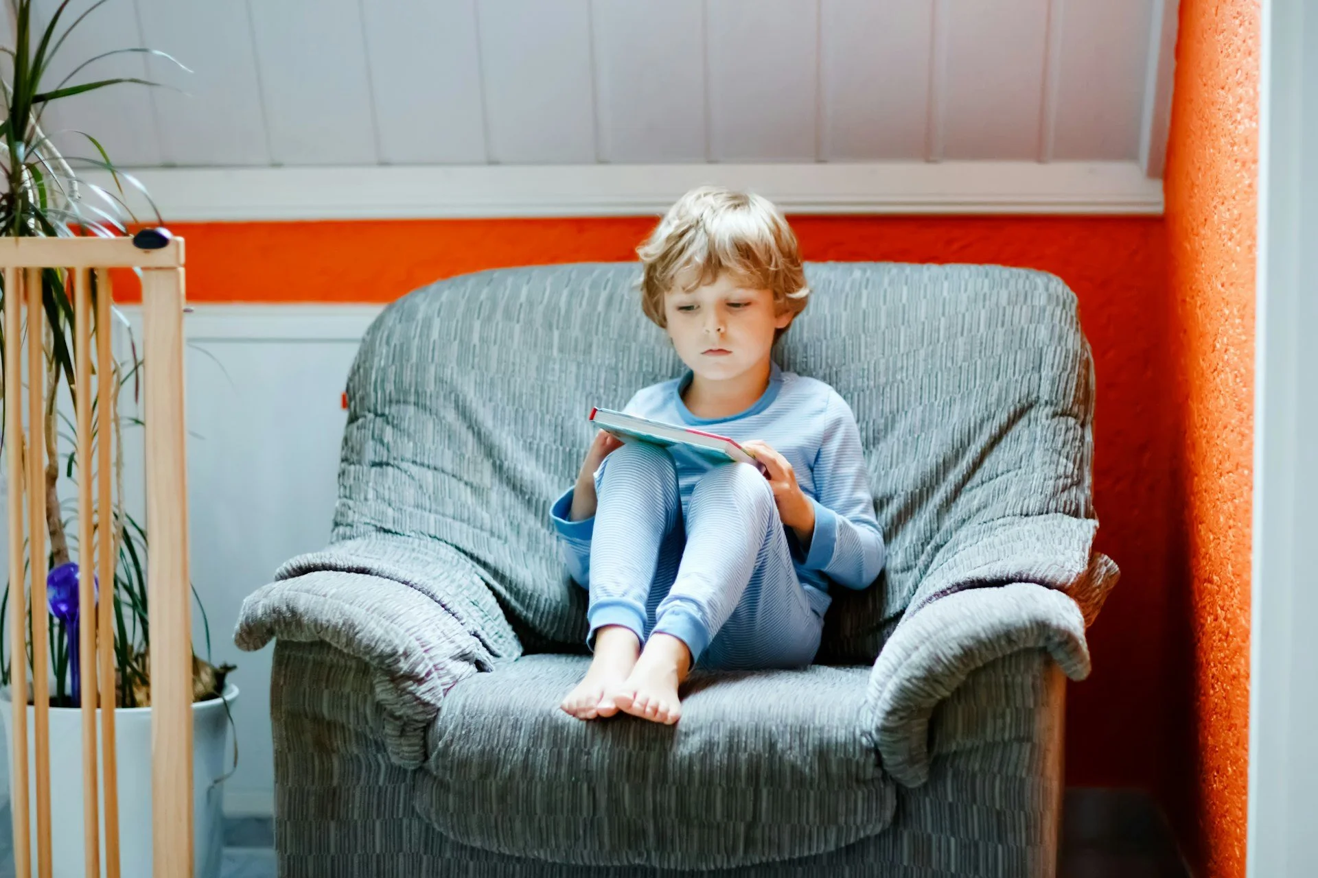 A young boy with blond hair and pajamas sitting on a cushioned armchair and reading a book, in a cozy room with an orange wall and a window.