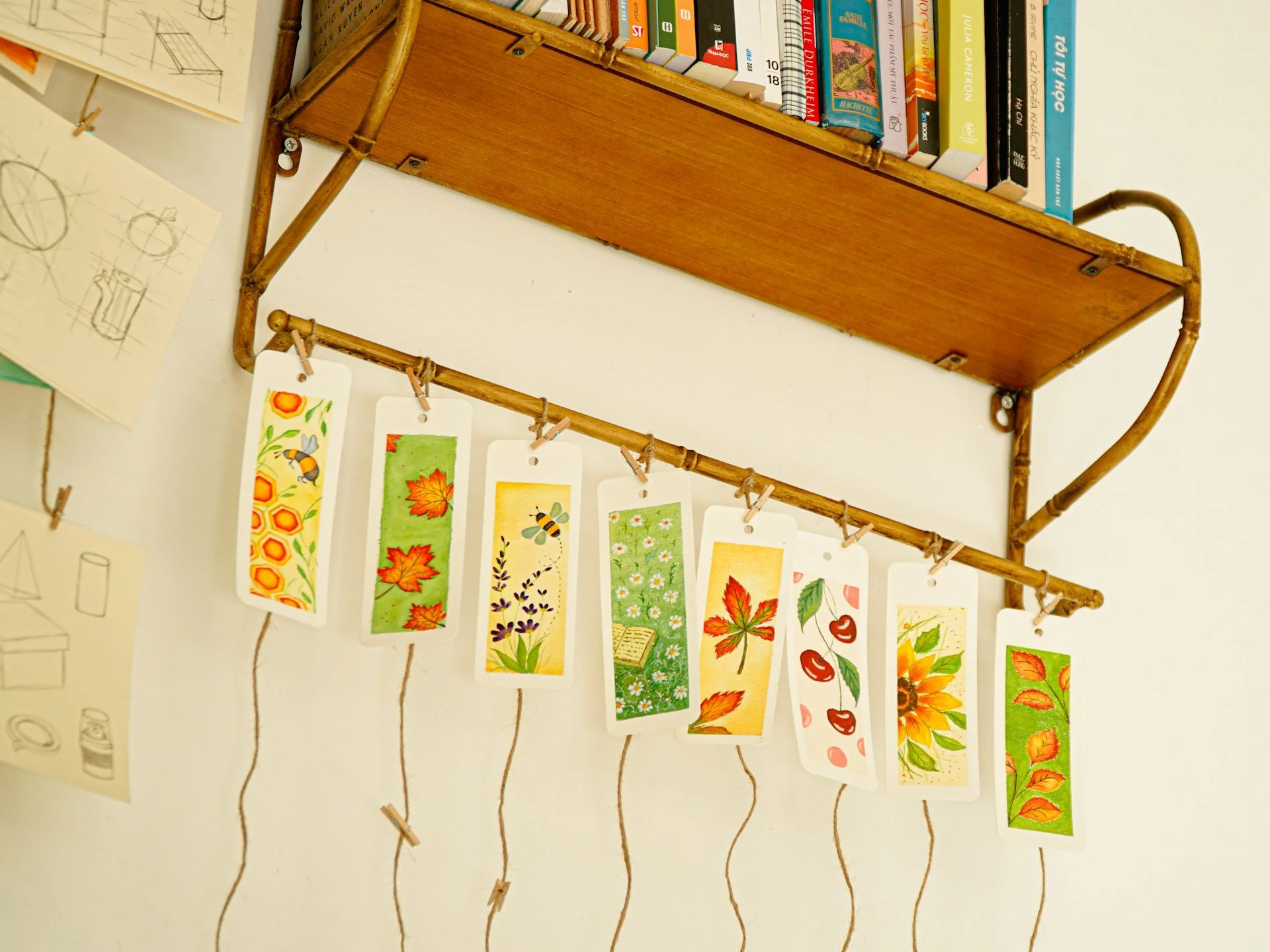 Wall-mounted display with illustrated bookmarks featuring colorful autumn themes such as leaves, bees, and flowers, hanging from a string with small wooden clothespins, alongside sketches and colorful books on a bookshelf.