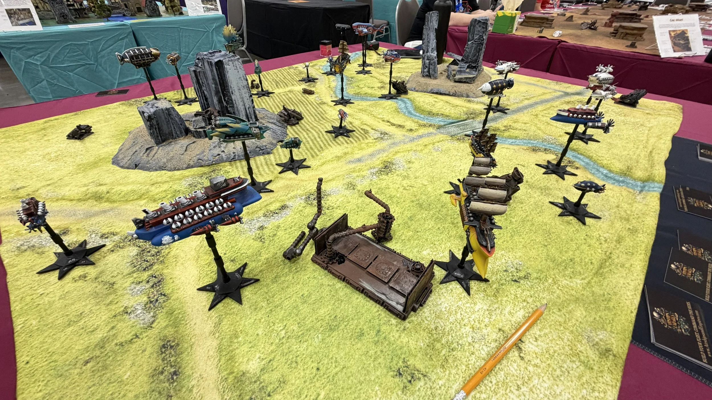 Tabletop miniature game with a detailed terrain, futuristic ships, and models resembling city ruins on a yellow surface at an exhibition or convention.
