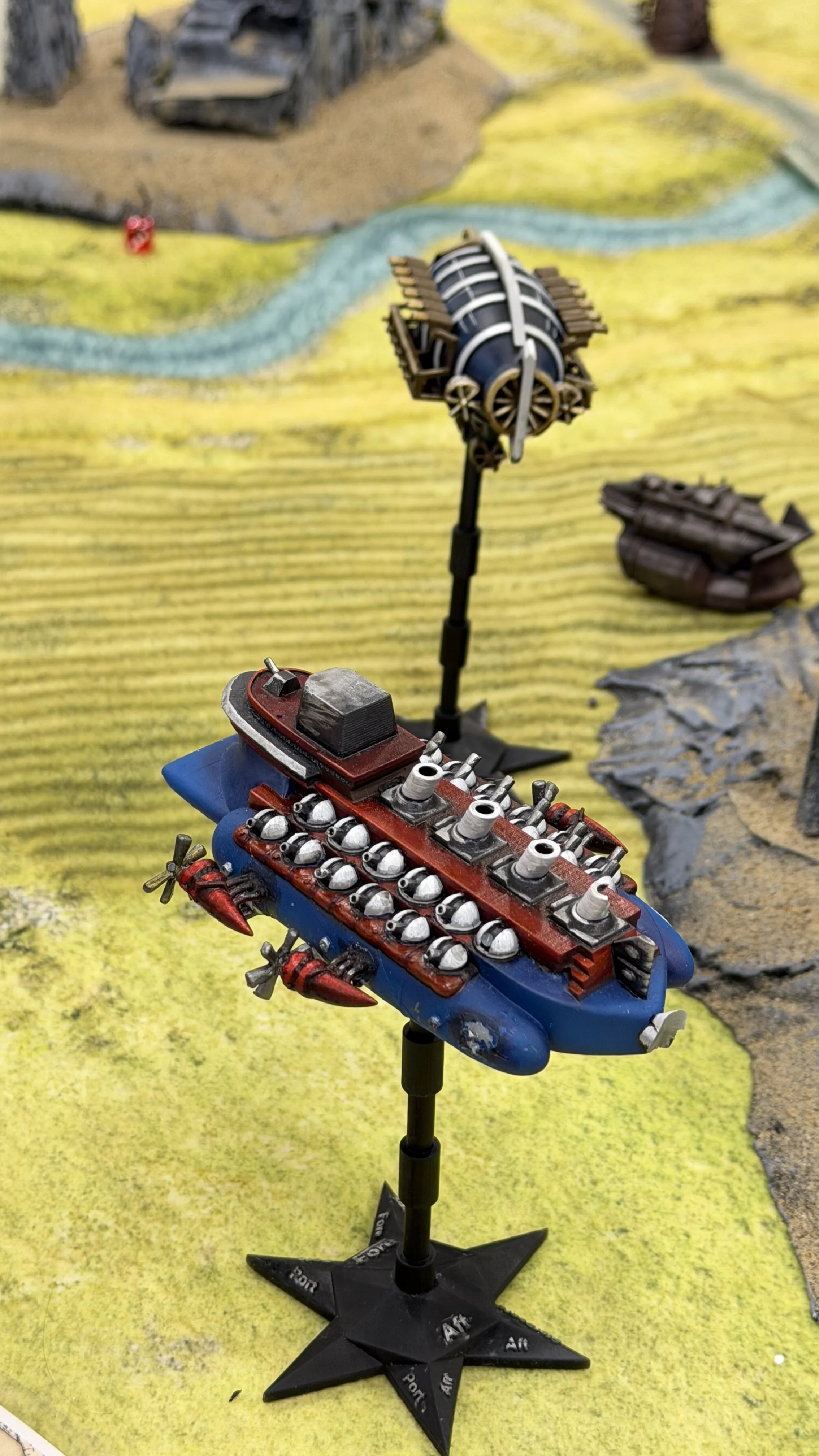 Miniature model of a red and blue battleship with numerous small laser cannons on top, mounted on a black star-shaped base, with a model of a helicopter in the background. The scene is set on a yellow textured surface with a painted landscape background featuring a river, rocky terrain, and a distant small red structure.