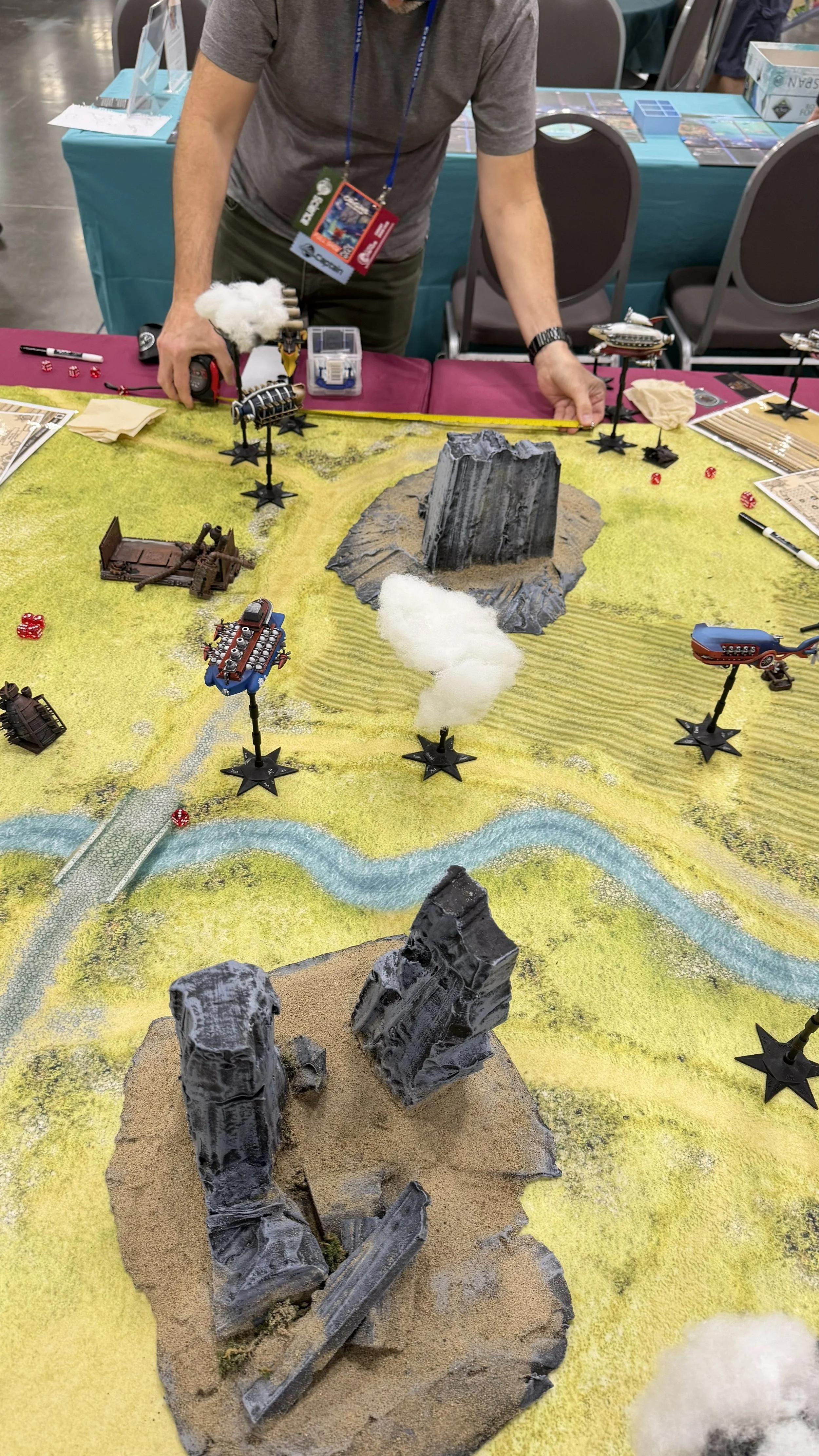 A tabletop game setup with terrain features, miniature vehicles, and structures. A person is adjusting the game pieces, which include a mountain, rocks, a river, and various models like ships, sci-fi aircraft, and buildings on a colorful game board.