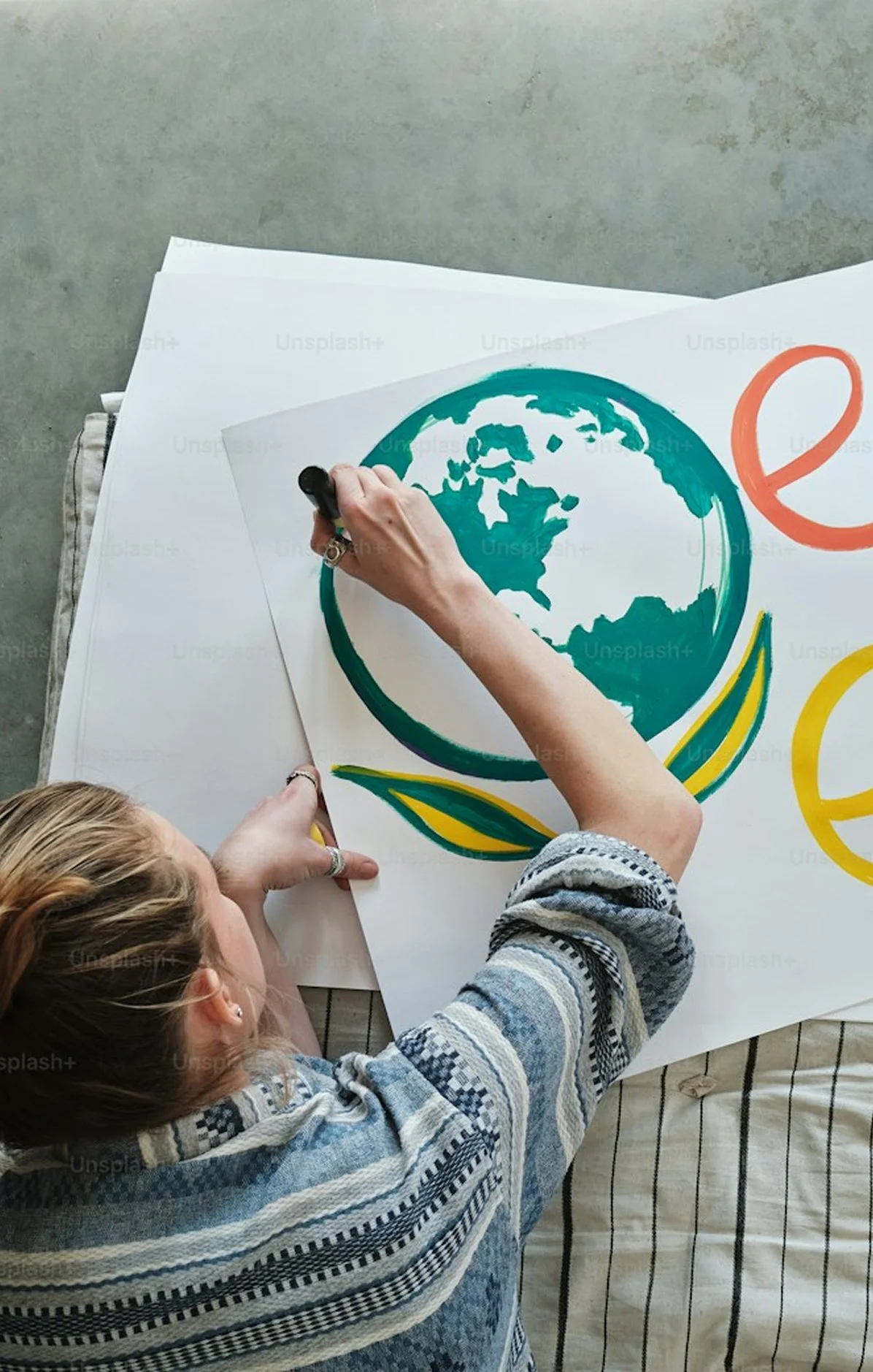 A person drawing on a large poster featuring a stylized Earth logo with green and blue colors, surrounded by abstract shapes and lines. The person is wearing a striped sweater and has long hair.