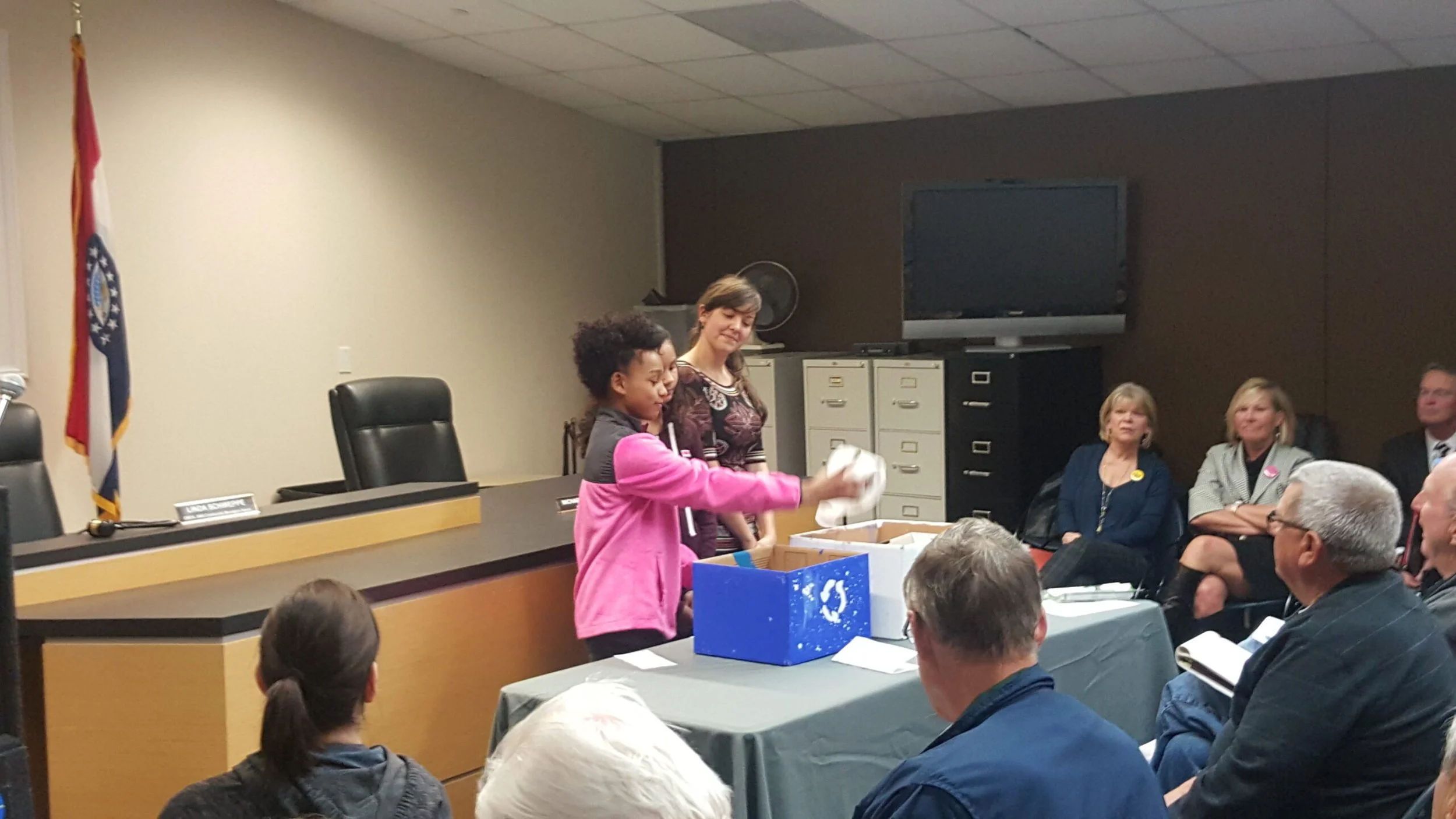 Young girl in pink jacket handing something to a woman in a meeting room with other adults seated around. There is a large blue box on the table and people are listening attentively.
