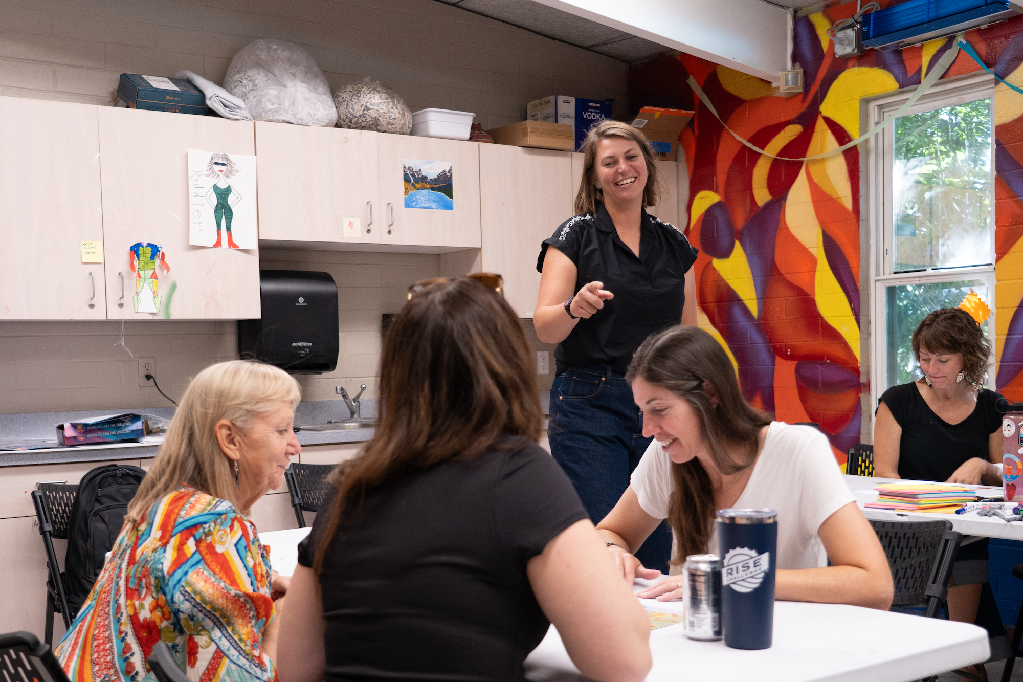A group of people sitting at tables in a room with colorful murals on the wall, while one person stands and gestures.