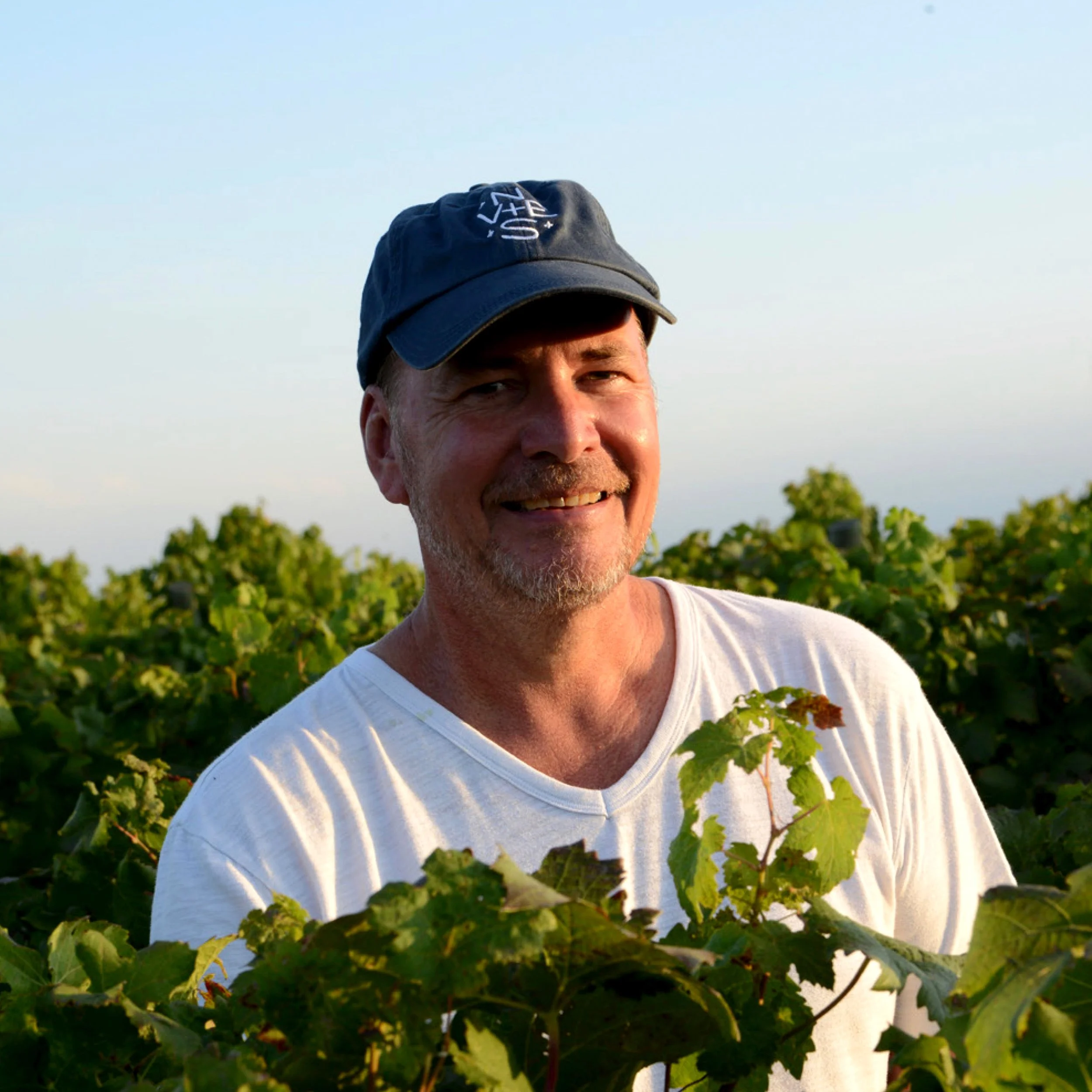 A smiling man outdoors wearing a navy blue cap and white t-shirt, surrounded by green vines and foliage.