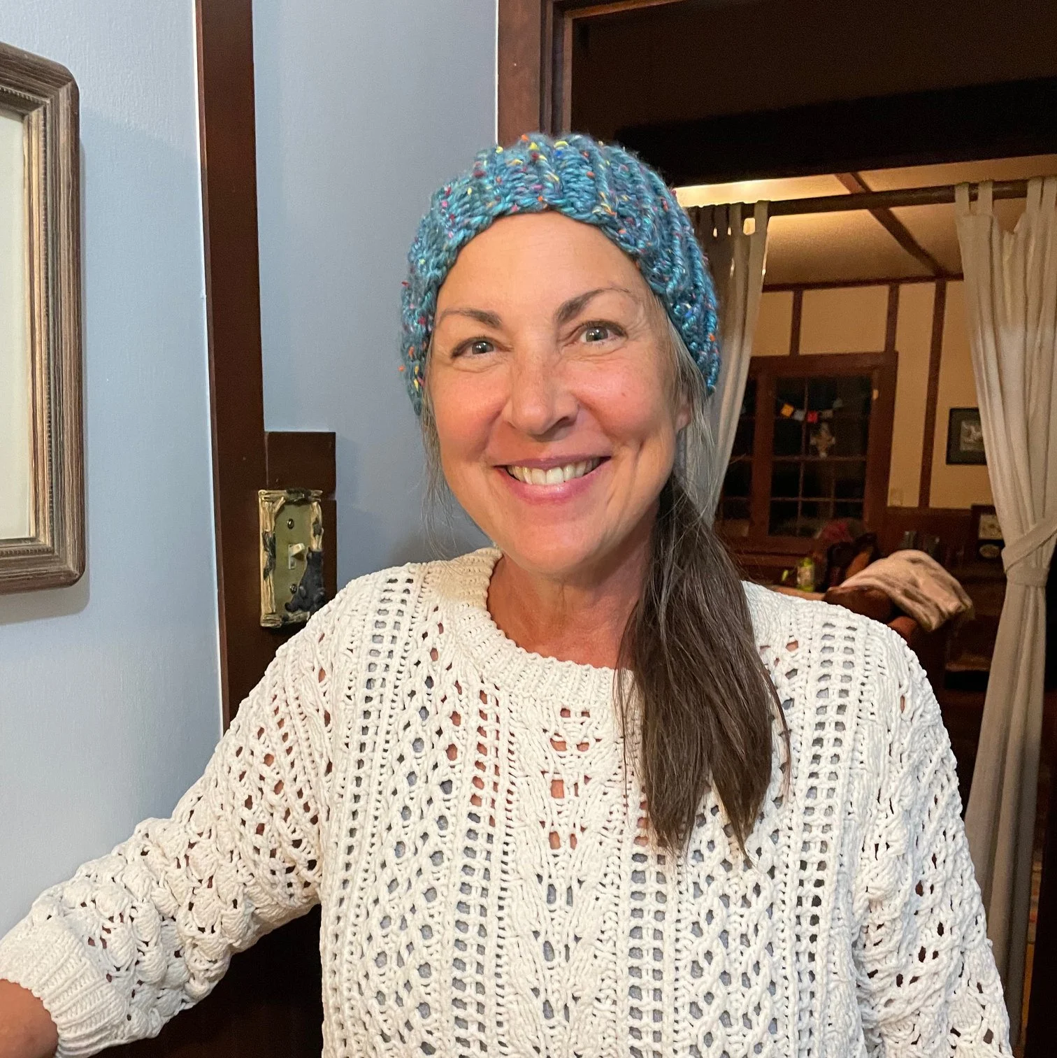 A woman with long brown hair is smiling, wearing a multicolored knitted headband and a white open-knit sweater, standing indoors in a warmly lit room.