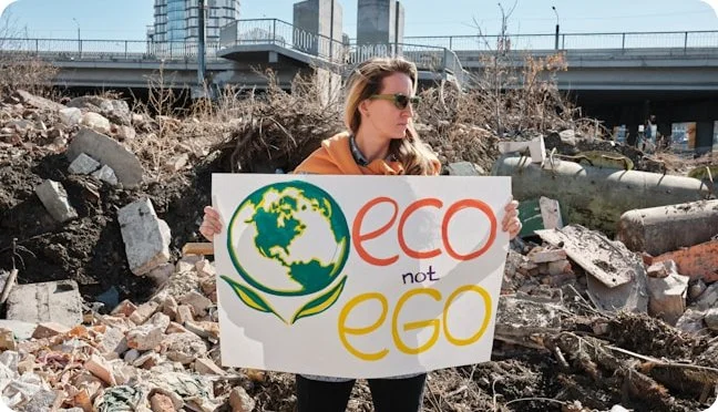 A woman wearing sunglasses and an orange jacket holding a sign that says "eco not ego" with a globe illustration, standing amidst debris and rubble under a bridge.