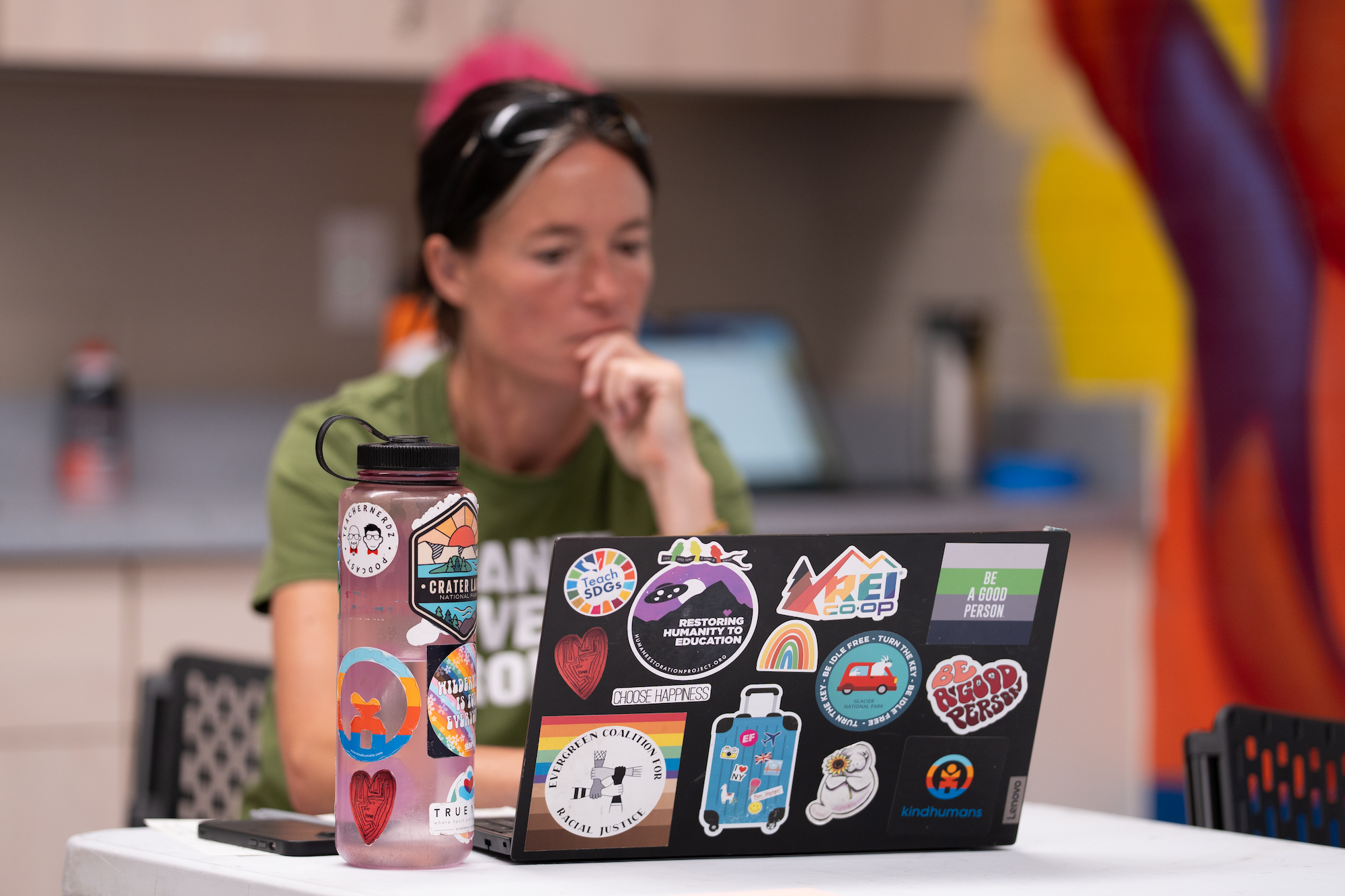 A person with their hand on their chin looks intently at a laptop covered in stickers. A water bottle also covered in stickers sits to the left of the laptop.