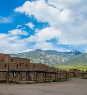 Old adobe buildings with wooden balconies in front of green mountains and a partly cloudy sky.