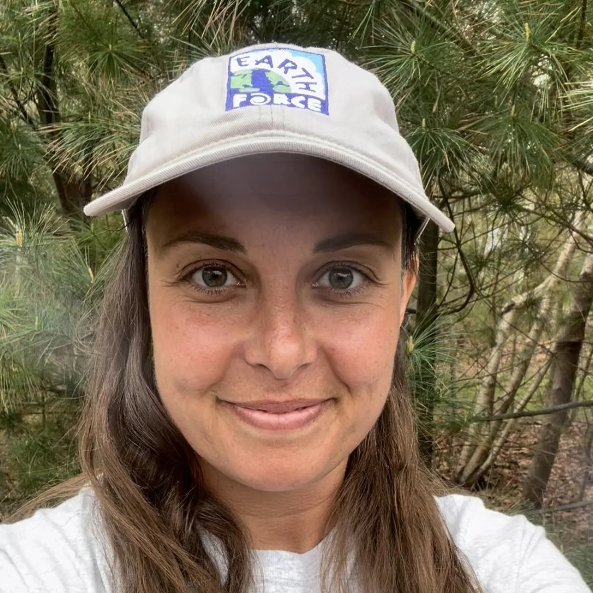 A woman wearing a gray hat with an Earth Force logo, outdoors with green trees in the background.