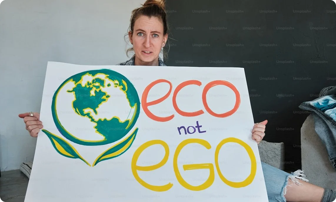 A woman holding a sign with a drawing of the Earth and the words 'eco not ego' in colorful letters.