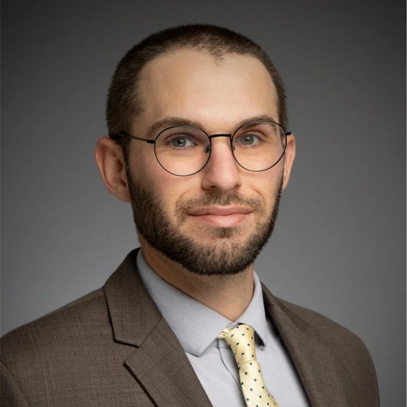 Professional portrait of a man with glasses, a beard, wearing a brown suit, light blue shirt, and yellow polka dot tie, against a gray background.