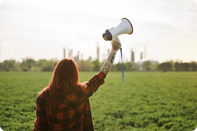 Person with red hair and tattoos holding a megaphone in a green field with modern city skyline in the background.