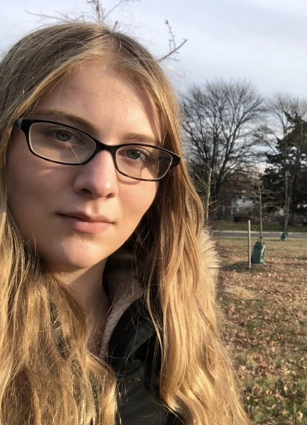 A young woman with long wavy blonde hair wearing glasses and a black jacket, outdoors in a park with bare trees and a partly cloudy sky.