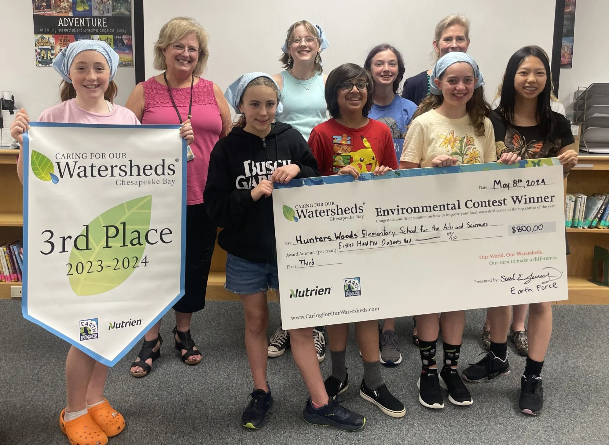 A group of children and two adults posing indoors, holding large award banners and a check for their environmental contest win. The banners display a third place award from Watersheds Chesapeake Bay for the school for the arts and sciences, dated May 8th, 2024. Some children are wearing headscarves, and everyone is smiling, celebrating their achievement.
