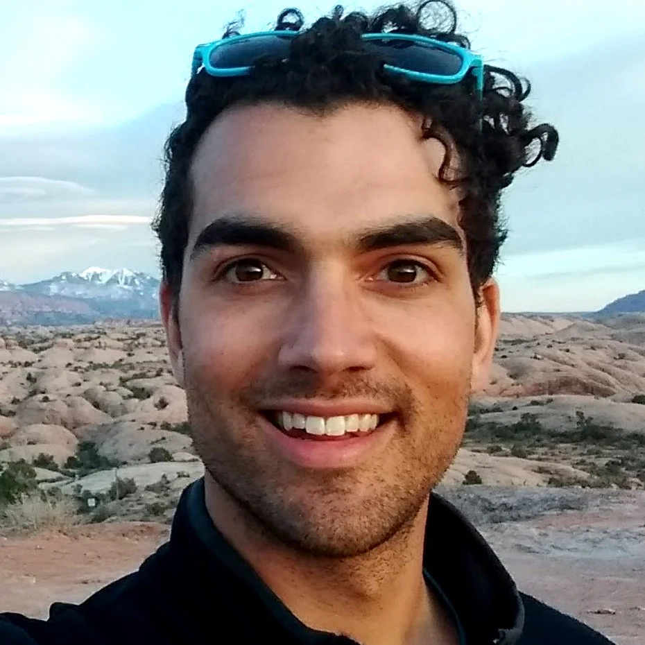 A man with curly dark hair smiling outdoors with mountains and rocky terrain in the background.