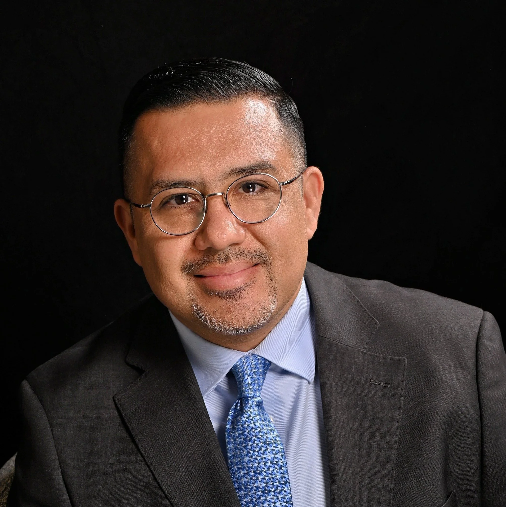A middle-aged man with short, dark hair wearing glasses, a dark gray suit, a light blue shirt, and a blue patterned tie, smiling against a black background.