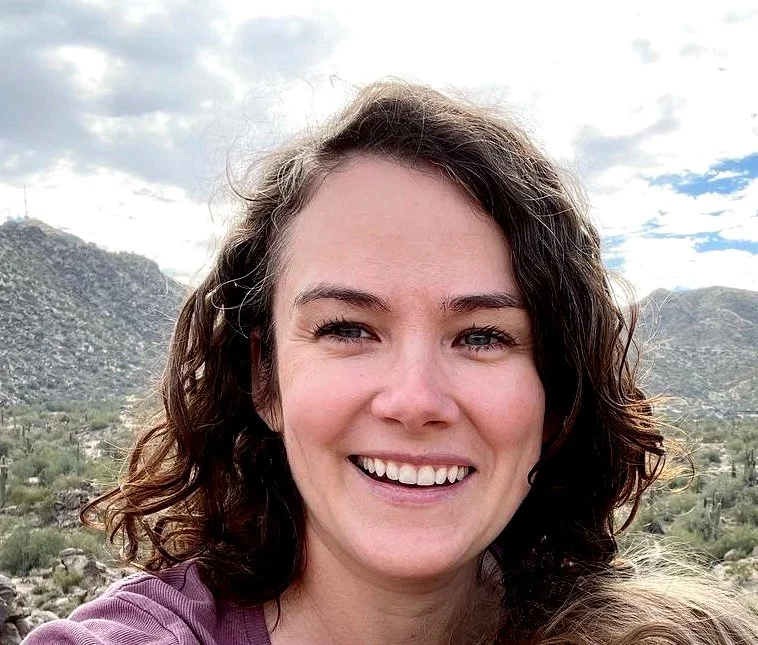 A woman with curly brown hair smiling outdoors with mountains and cloudy sky in the background.