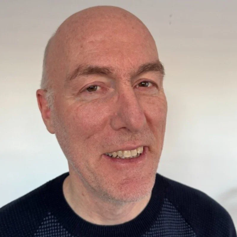 Close-up of a middle-aged man with a bald head, slight stubble, and wearing a black shirt, smiling with visible teeth against a neutral background.