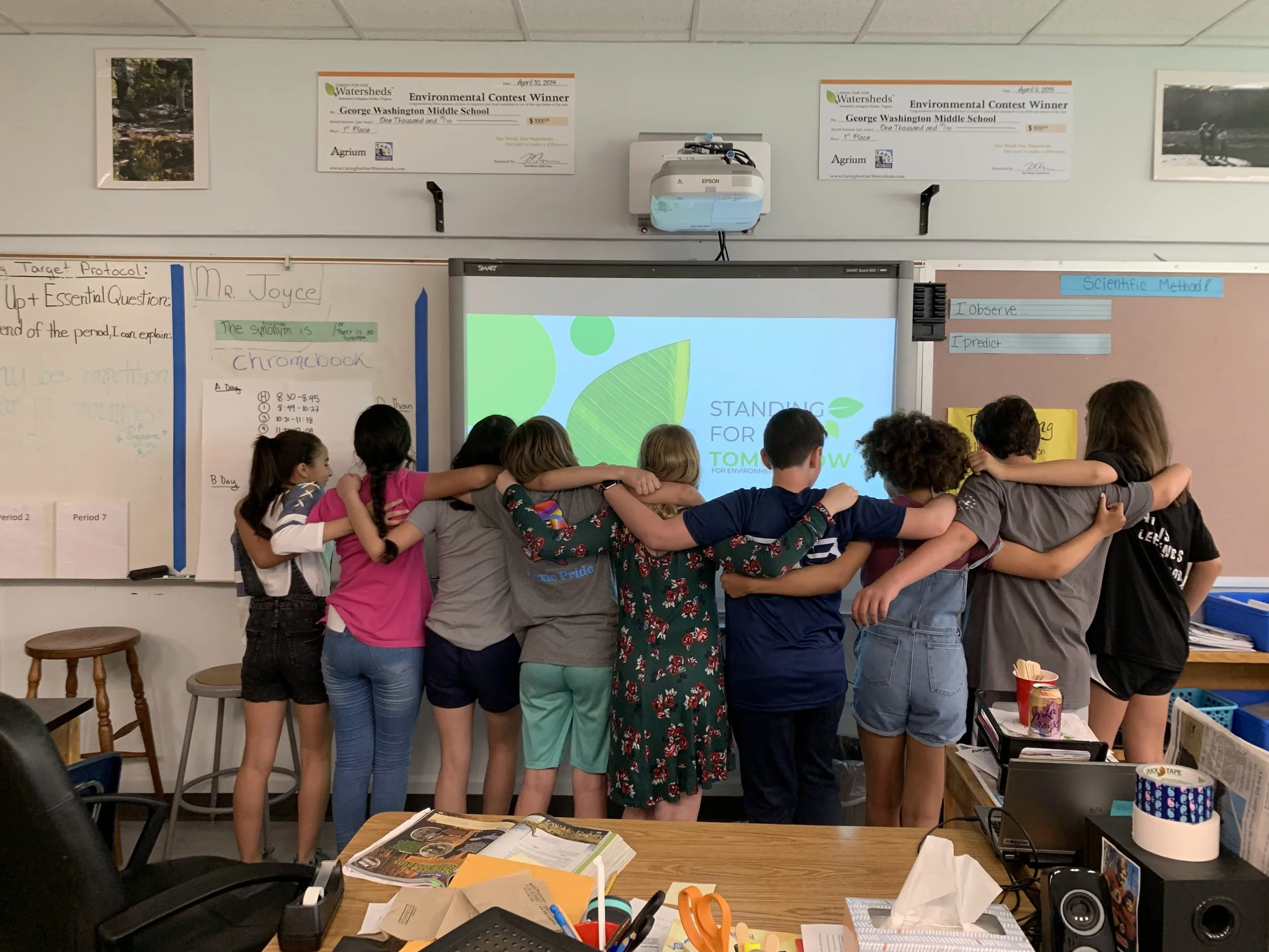 A group of children hugging in a classroom, standing in front of a whiteboard with educational posters, a smart board displaying a presentation, and two large checks on the wall.