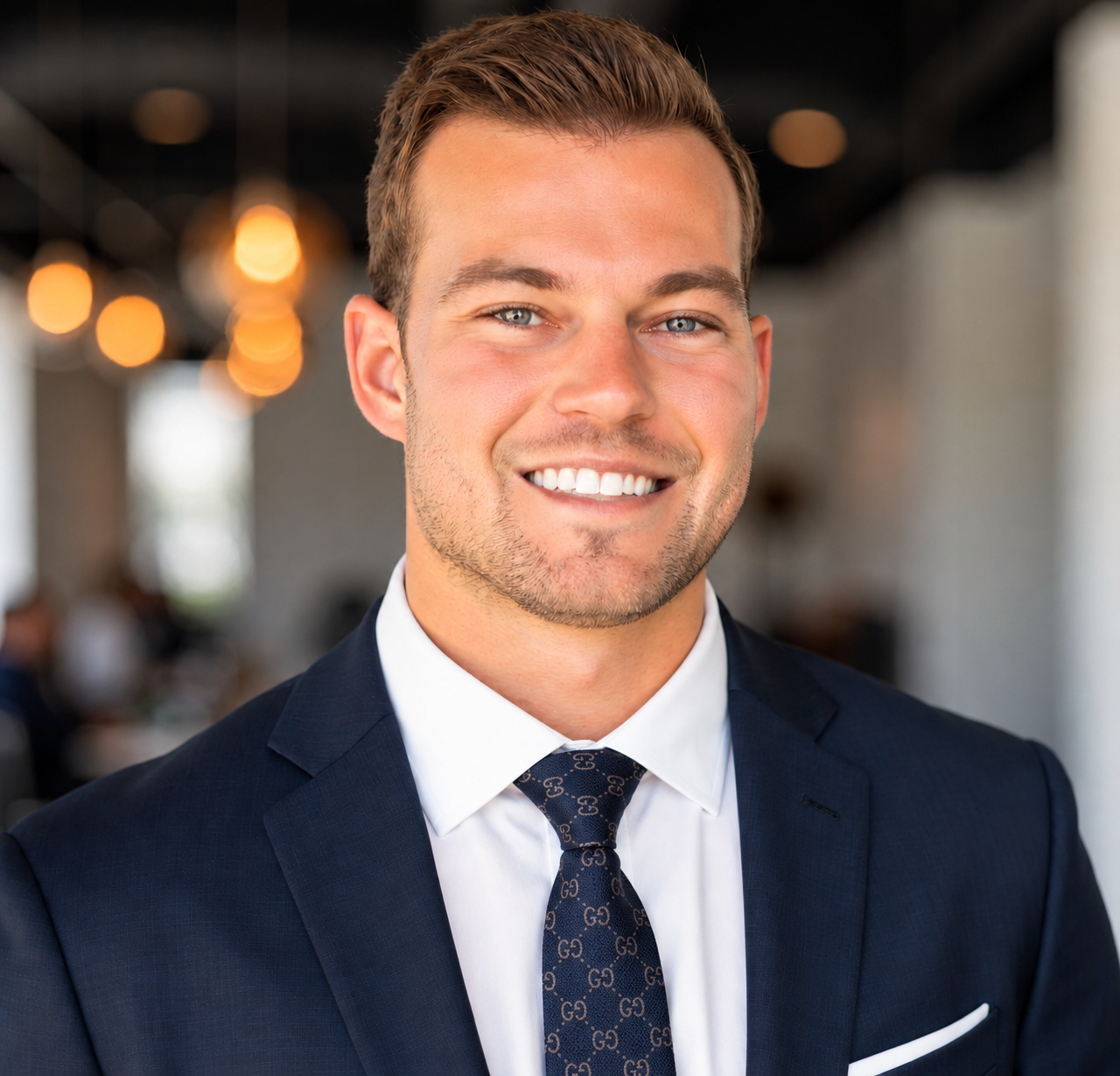 A smiling young man with short brown hair, blue eyes, and light skin wearing a navy suit, white shirt, and a patterned navy tie in an indoor setting.