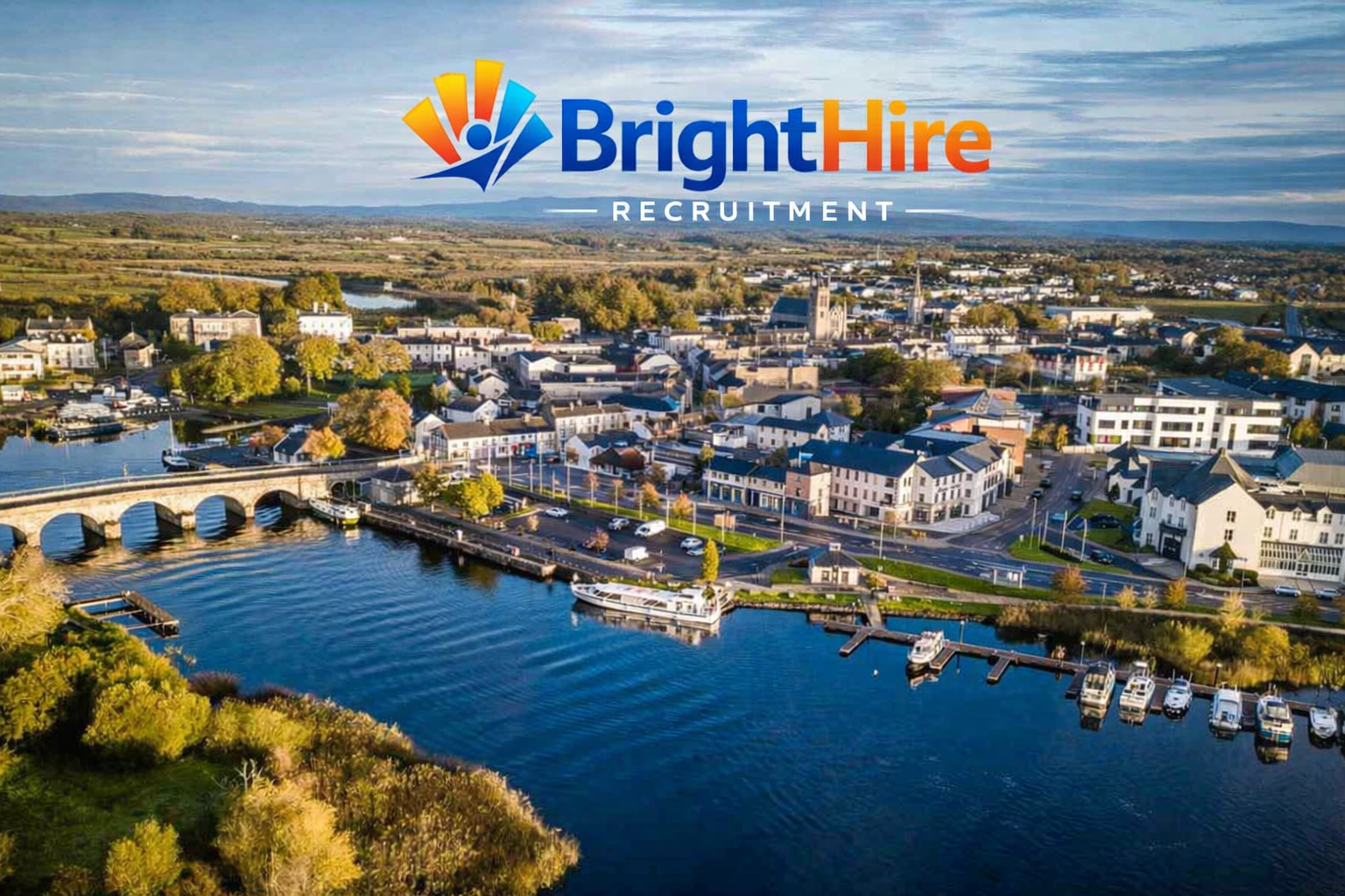 An aerial view of a small town with a river, boats, a stone bridge, and a mix of residential and commercial buildings, with trees displaying fall colors and a partly cloudy sky.