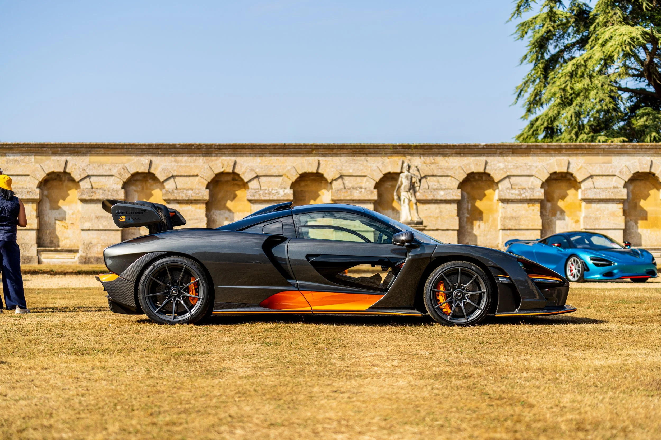 A black and orange McLaren sports car parked on grass with a yellow sports car and a blue sports car behind it, set against an old stone wall with arches and a statue, under a clear blue sky.