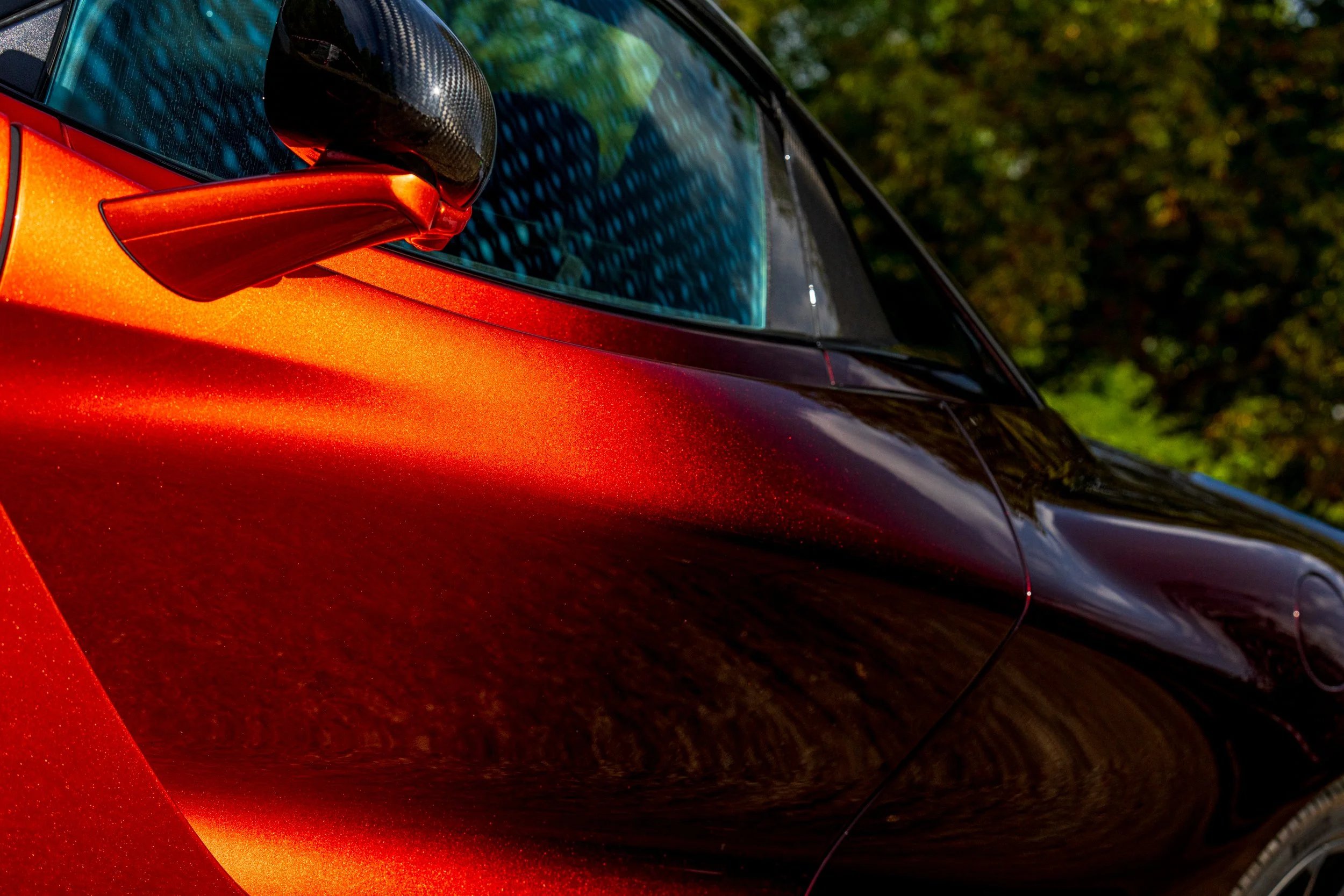 Close-up of a red sports car's side mirror and window, with a background of green trees.