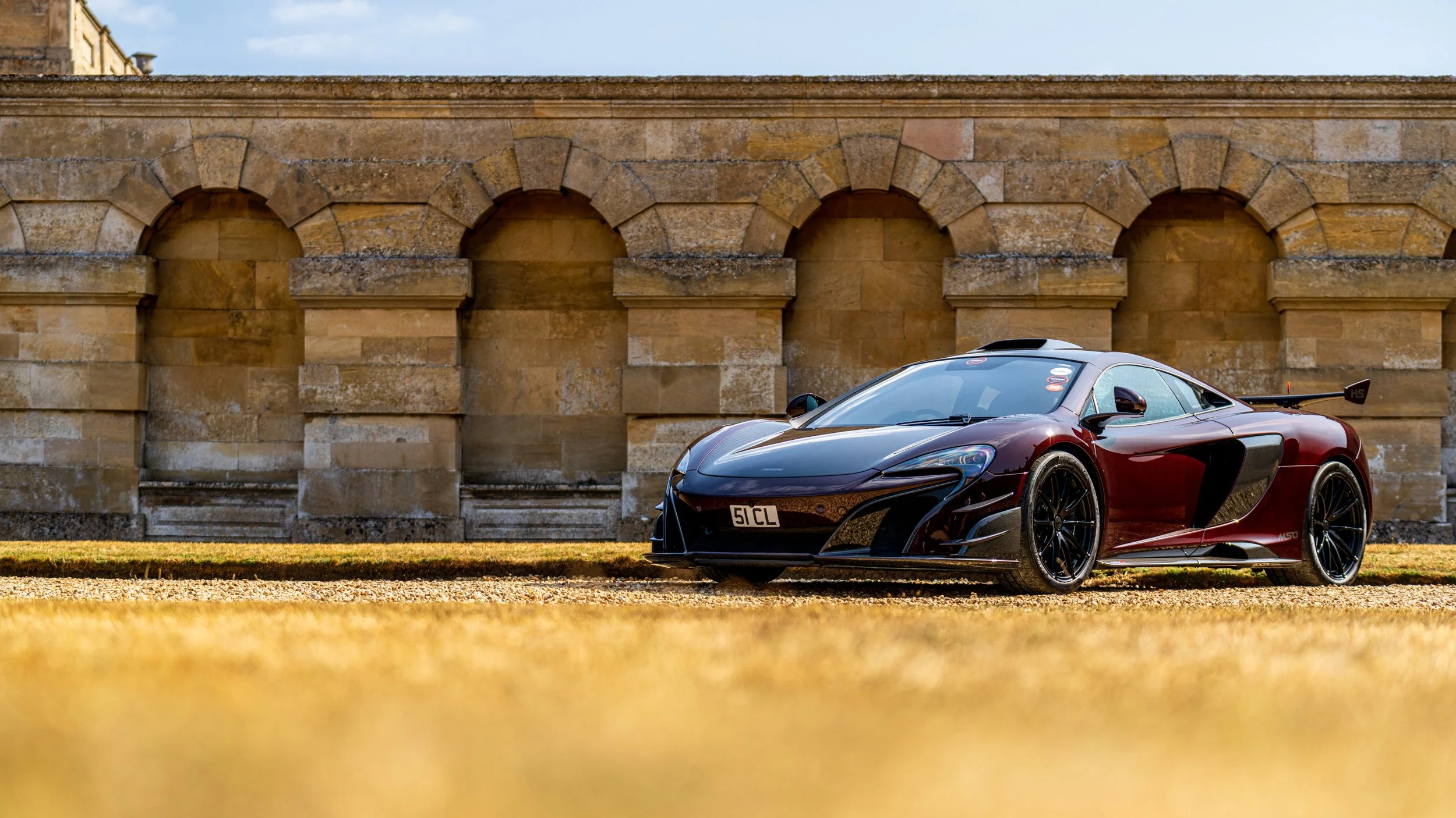 A luxury sports car parked in front of an old stone wall with arched openings under a blue sky.