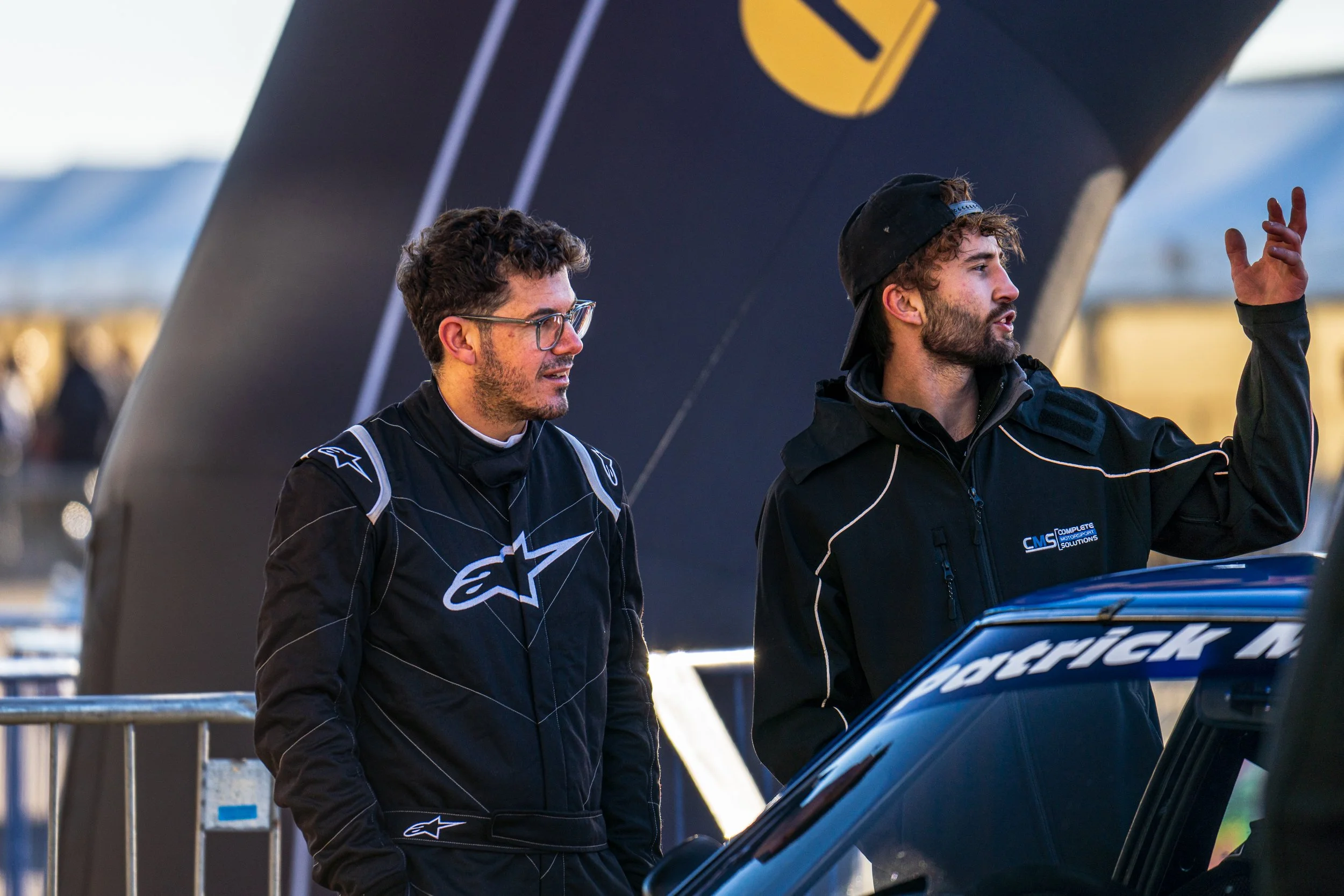 Two team members in dark clothing beside a racing car in the Silverstone paddock.